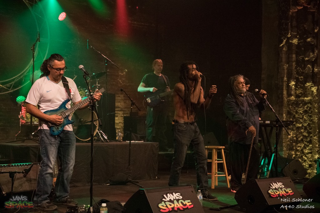Four musicians perform on stage with colorful lighting, one playing guitar, others singing and playing instruments, brick wall background.