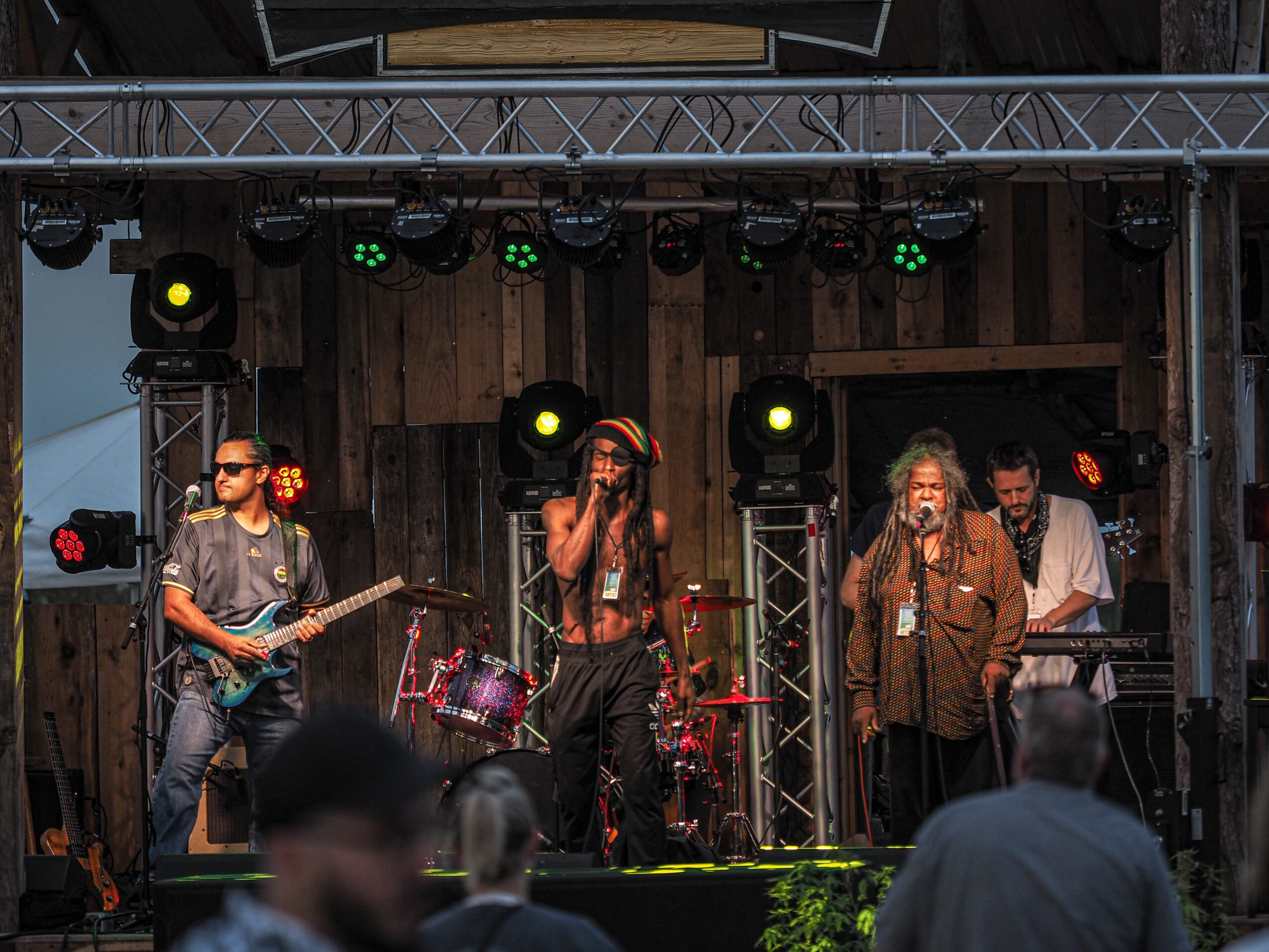 Band performing on stage with four musicians, wooden backdrop, stage lights, and audience in foreground.