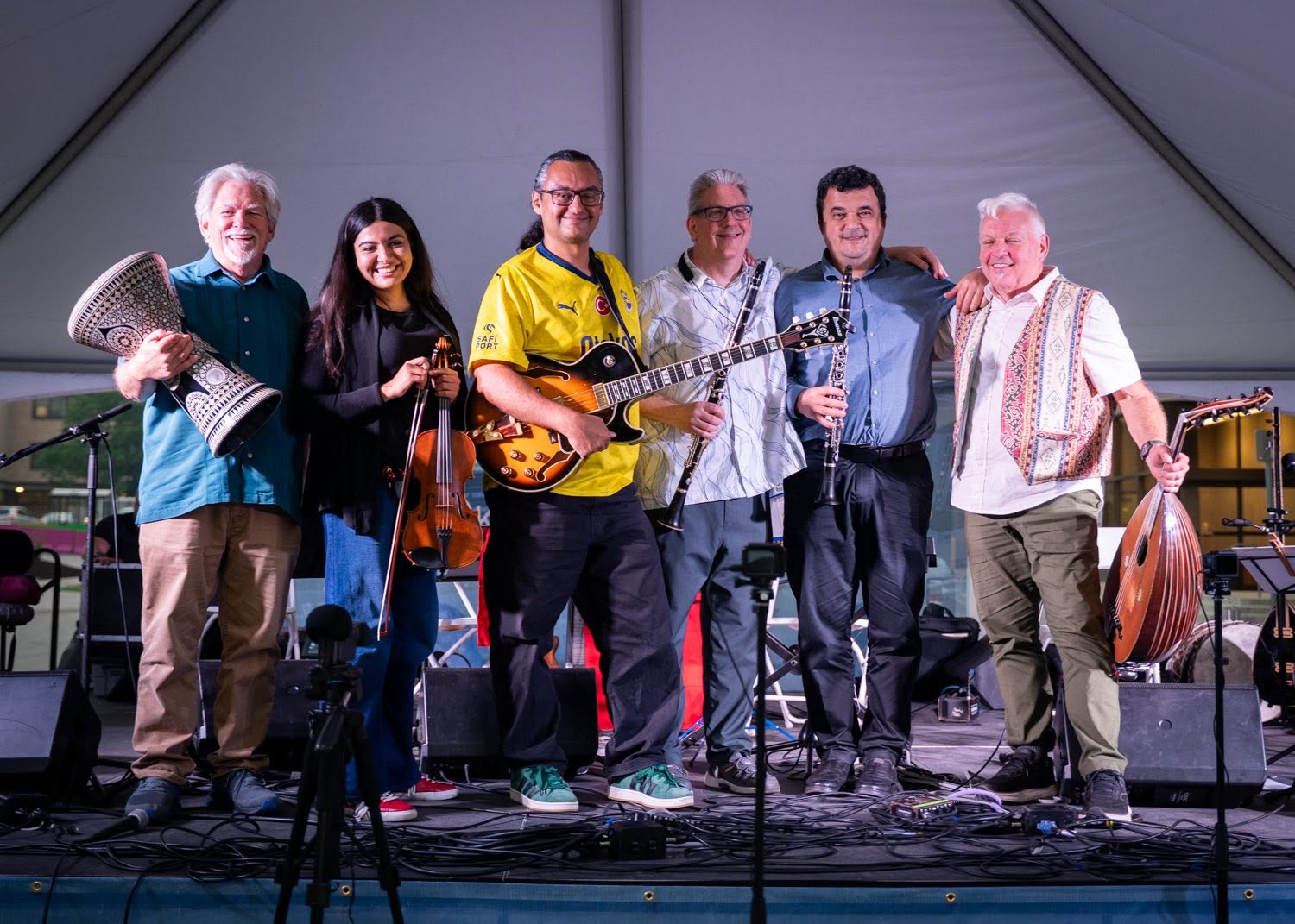 Six people standing on a stage with musical instruments, smiling, in a large indoor space.