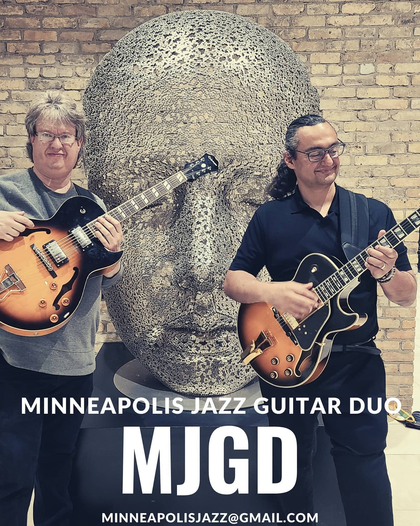 Two musicians with guitars stand in front of a large stone head sculpture against a brick wall. Text reads 'MINNEAPOLIS JAZZ GUITAR DUO MIGD'.