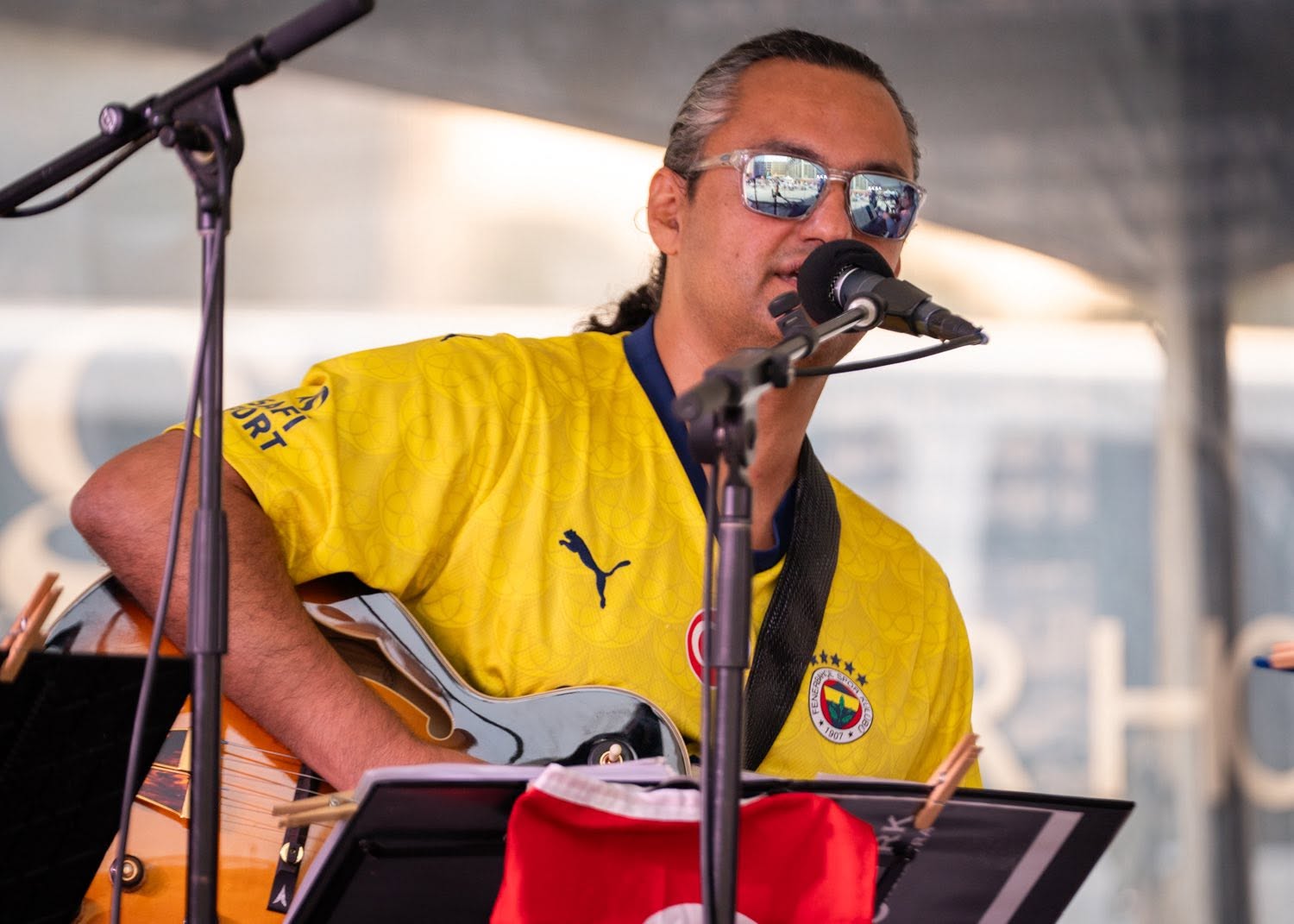 Person wearing yellow shirt playing guitar and singing into microphone at an outdoor event.