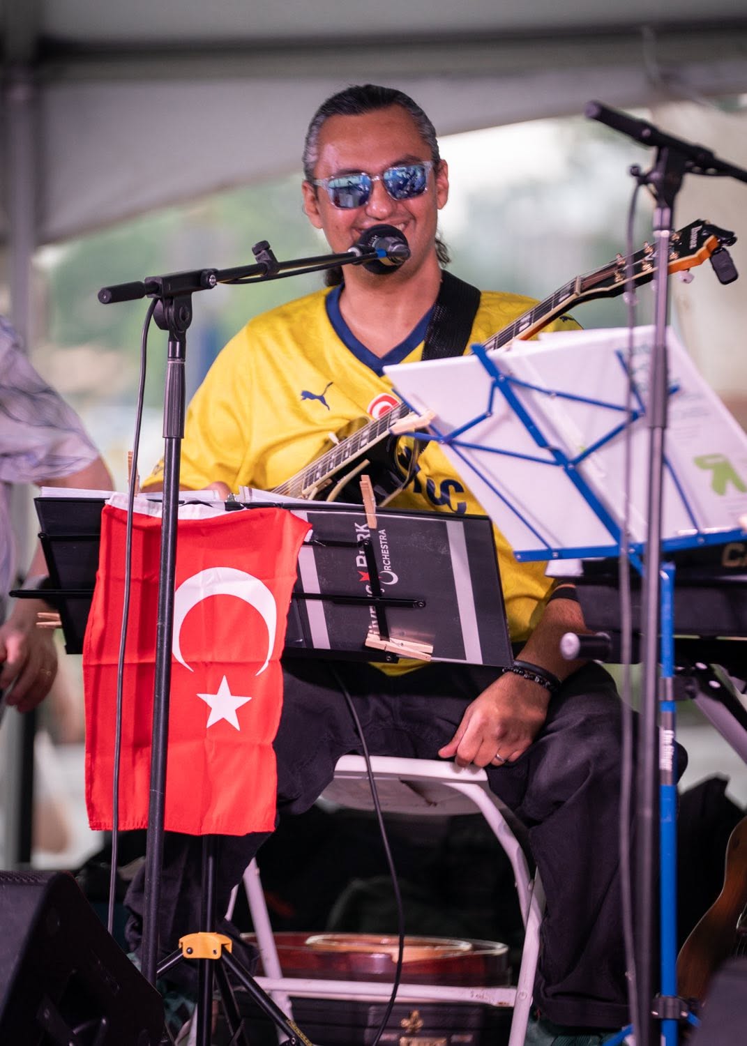 Man wearing sunglasses and a yellow sports jersey, sitting with musical equipment and a Turkish flag, under a tent.