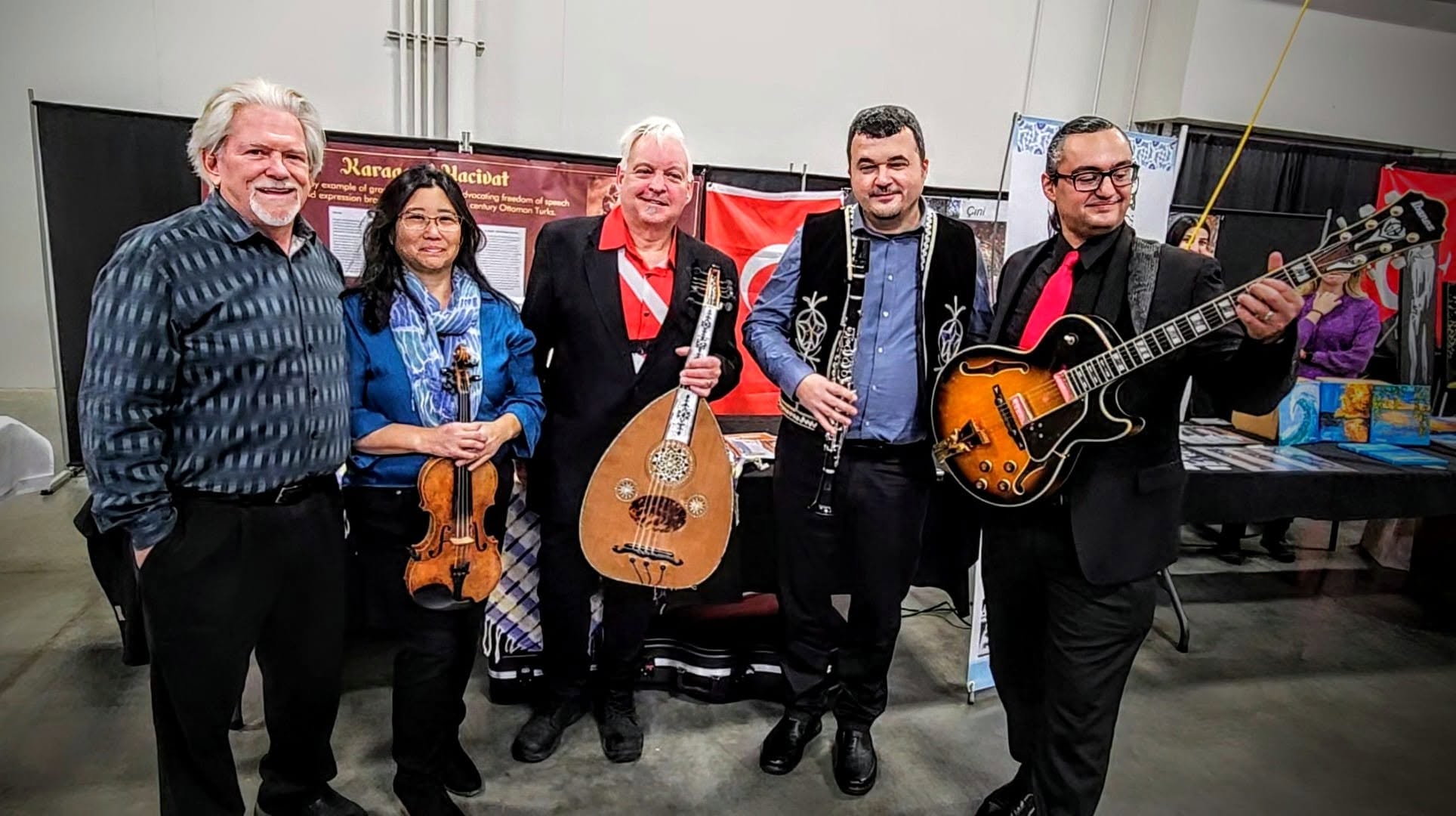 Five people standing together, some holding musical instruments, in an indoor setting with colorful background elements.