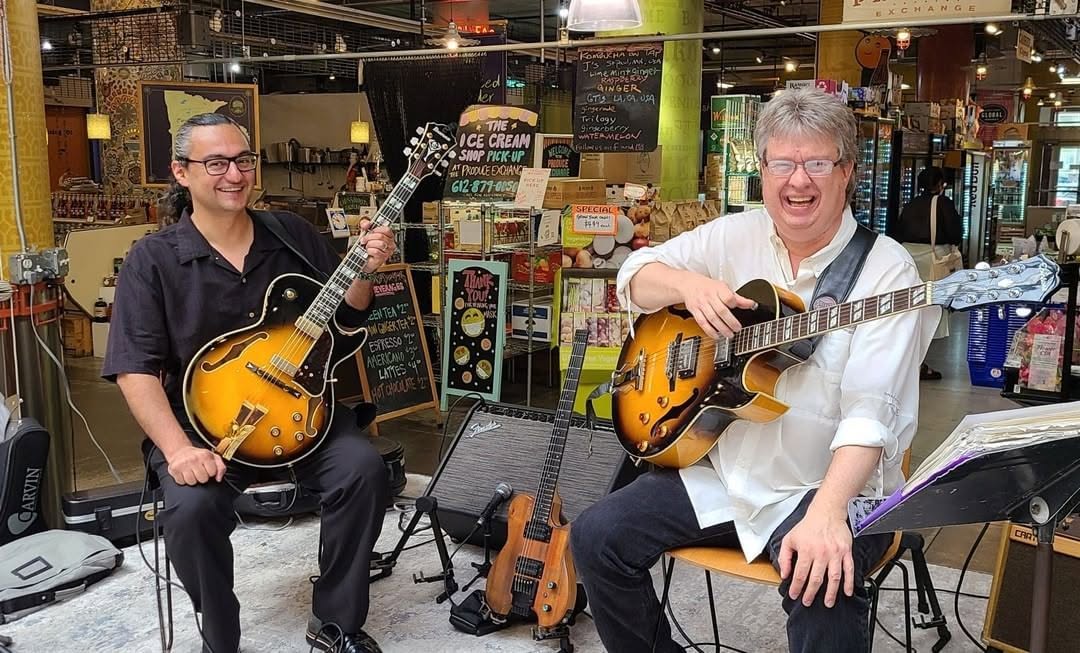 Two men playing guitars in a store, smiling, with music equipment and shelves in the background.