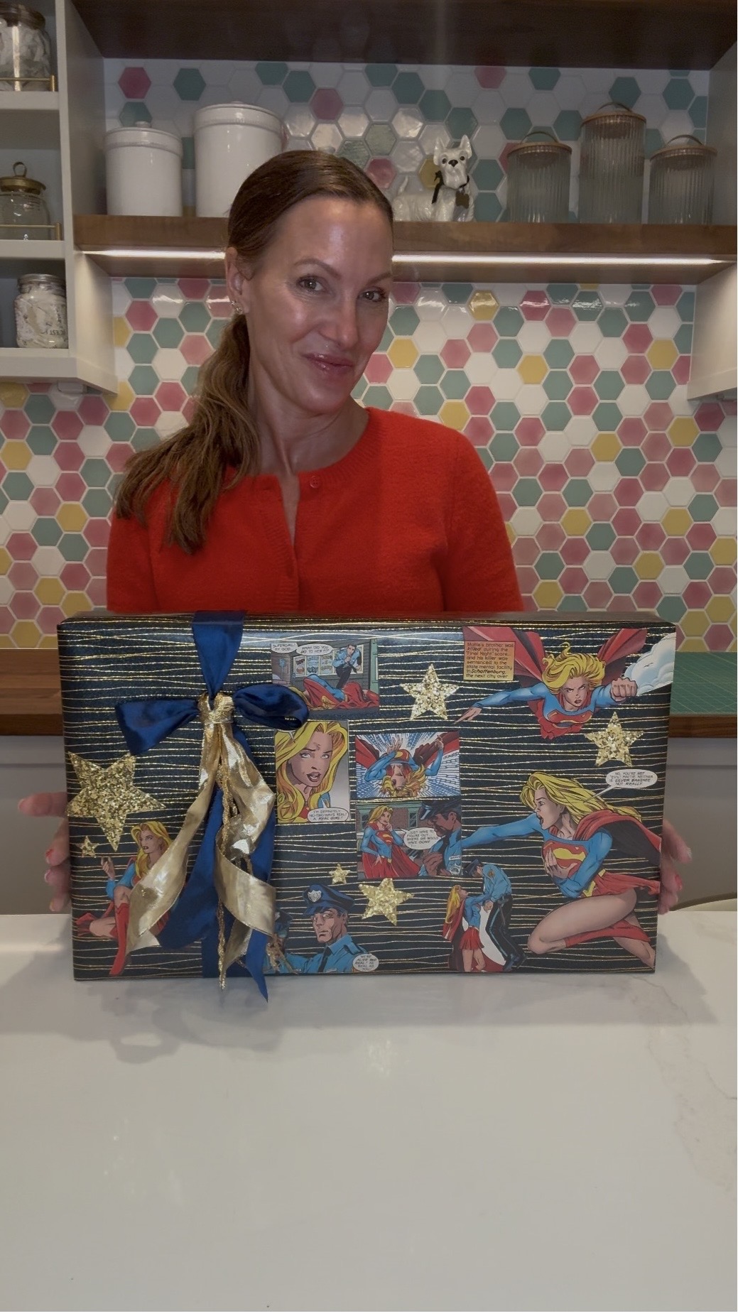 Woman with red hair and red top behind a decorated box, colorful tiled wall in background.