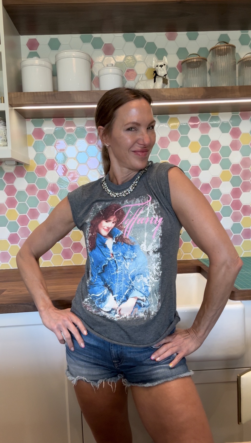 Woman with red hair wearing a graphic T-shirt and denim shorts, standing in front of a colorful tiled wall.