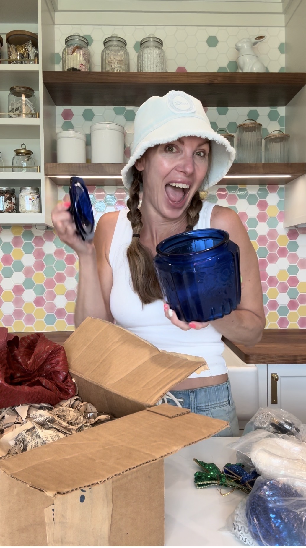 Woman wearing a white hat holding a blue container, smiling and excited in a kitchen with colorful tiles.
