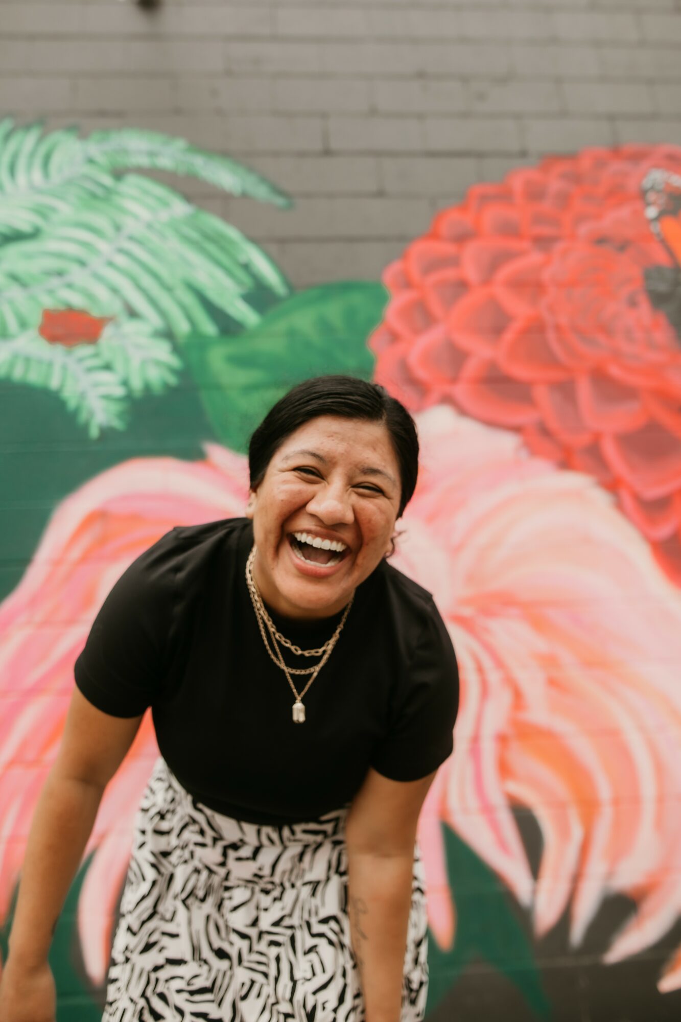 Person smiling and laughing, wearing a black top and patterned skirt, standing in front of a colorful floral mural.