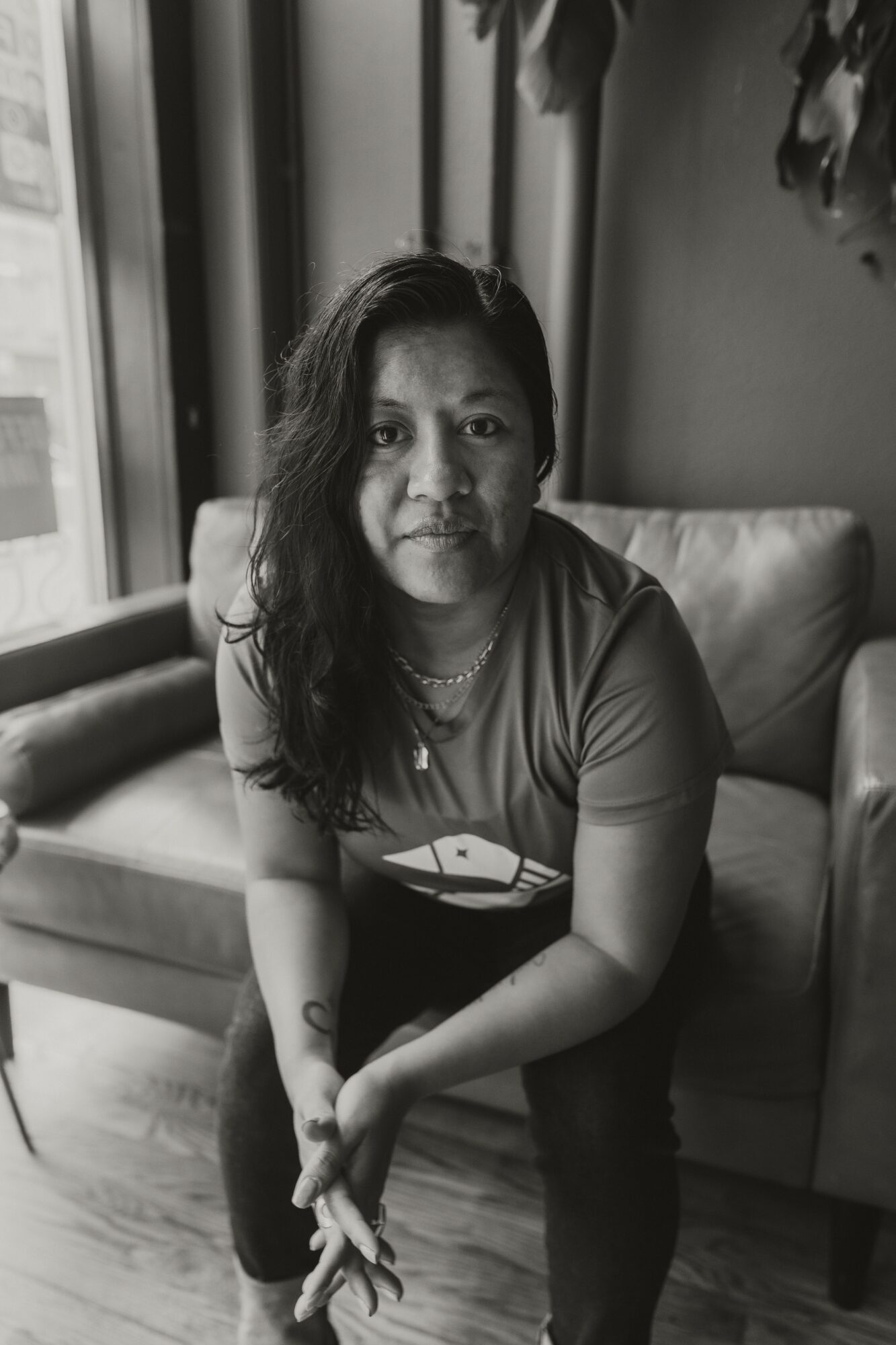 Woman with long hair sitting on a sofa indoors, looking at the camera, in black and white.
