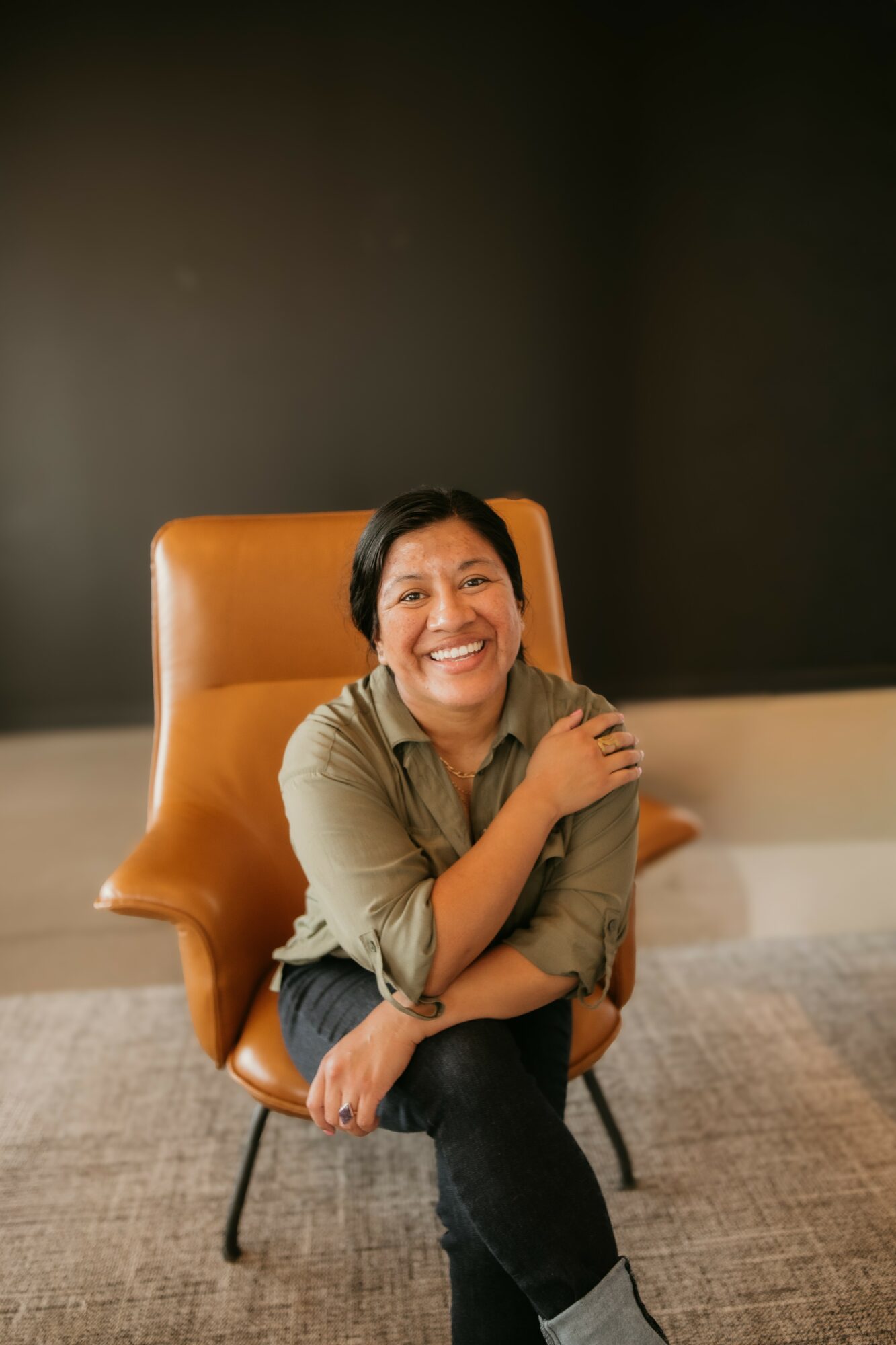 Smiling woman sitting on an orange chair with a dark background.