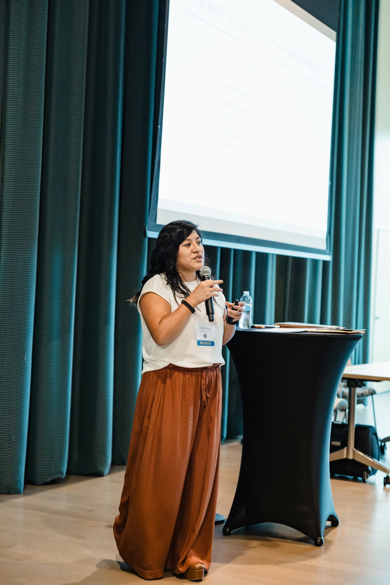 Woman standing at a podium, speaking into a microphone in a conference room with teal curtains and a large screen behind her.