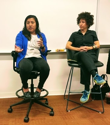 Two women seated and speaking in a room with a whiteboard behind them.