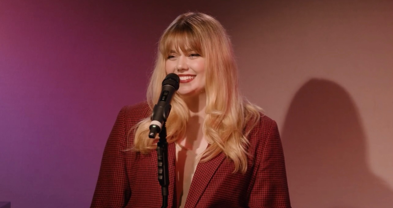 Woman with long blonde hair smiling at microphone on stage, background with shadow, wearing a dark red blazer.