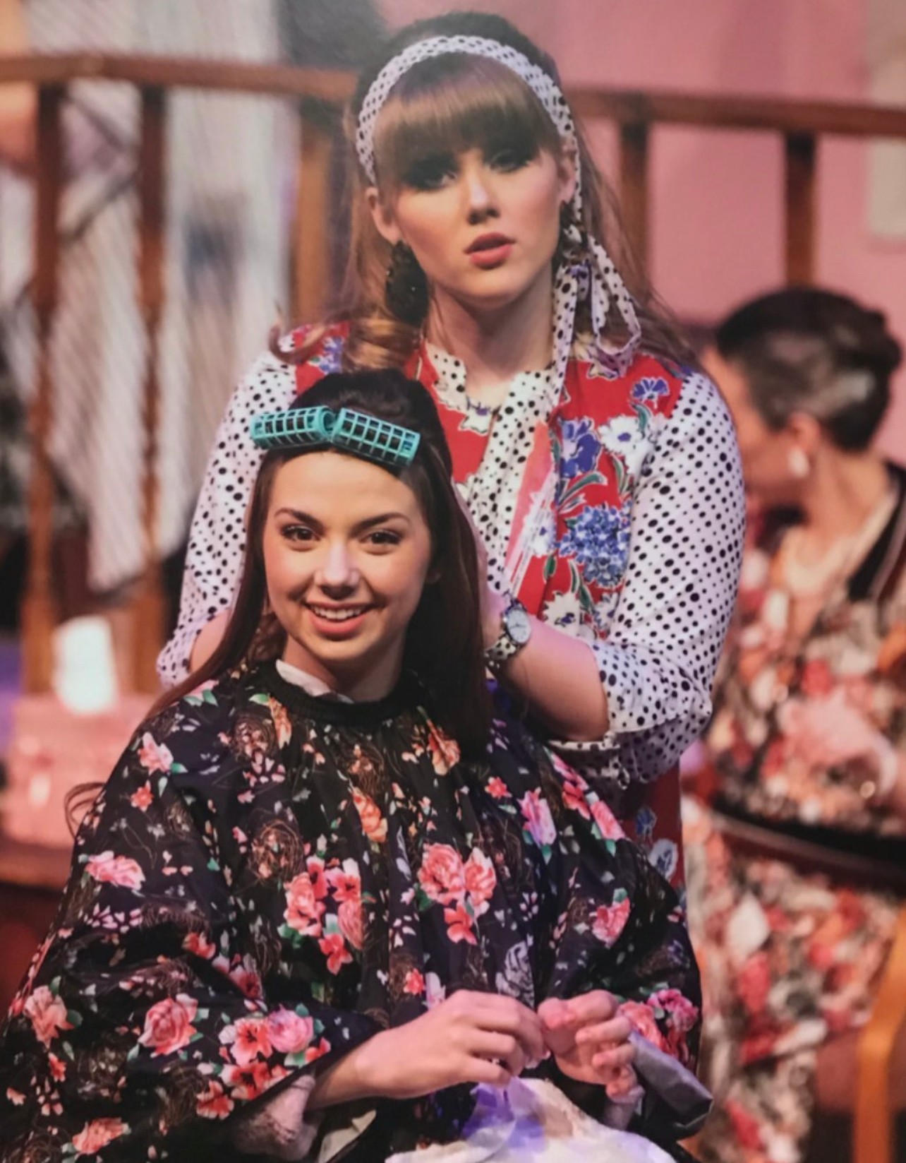 Two women, one smiling and seated, the other standing behind, in colorful, patterned clothing, indoors with a pink background.