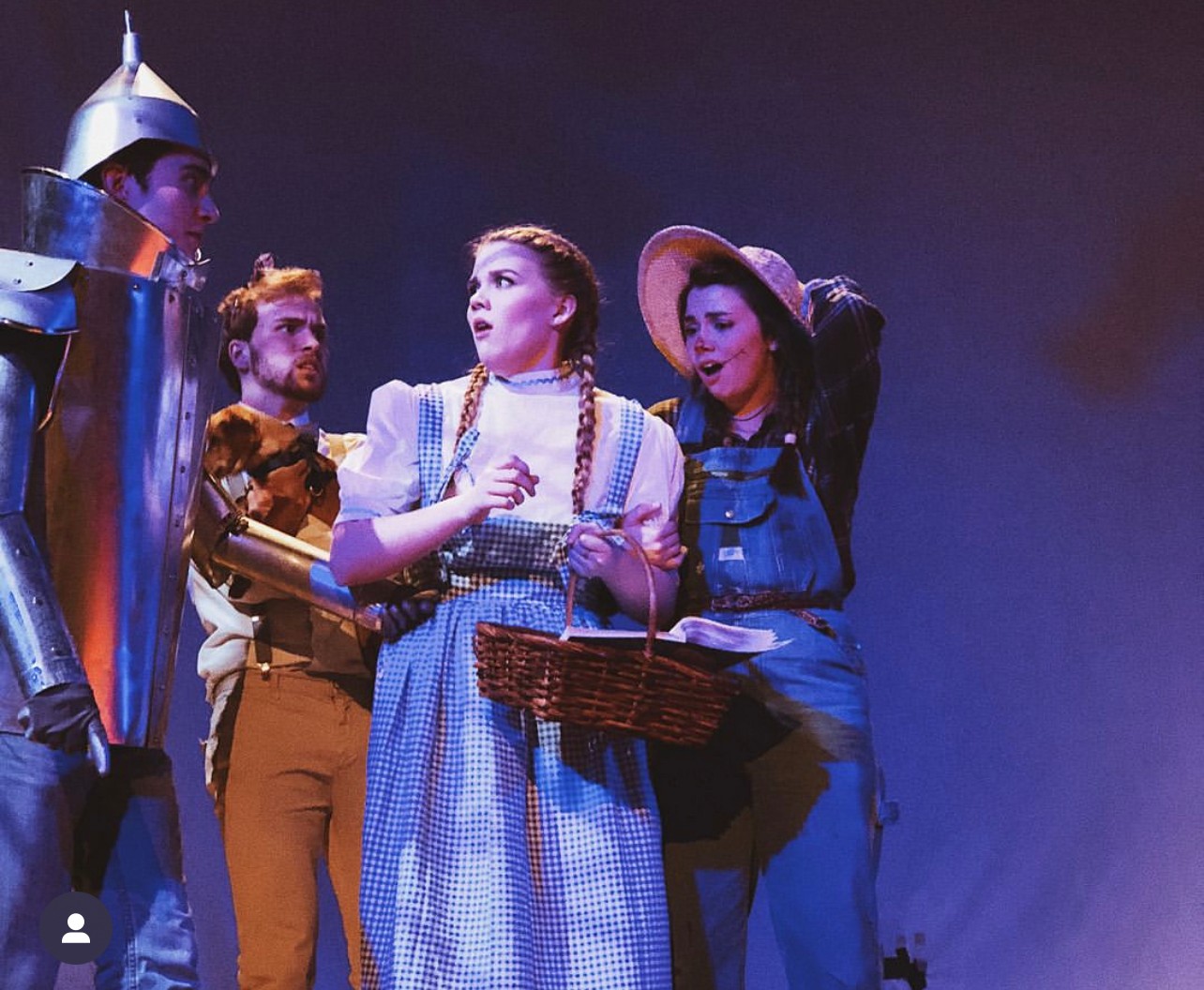 Four people in costume on stage, with a dark background, engaged in a scene, one holding a basket, others listening.