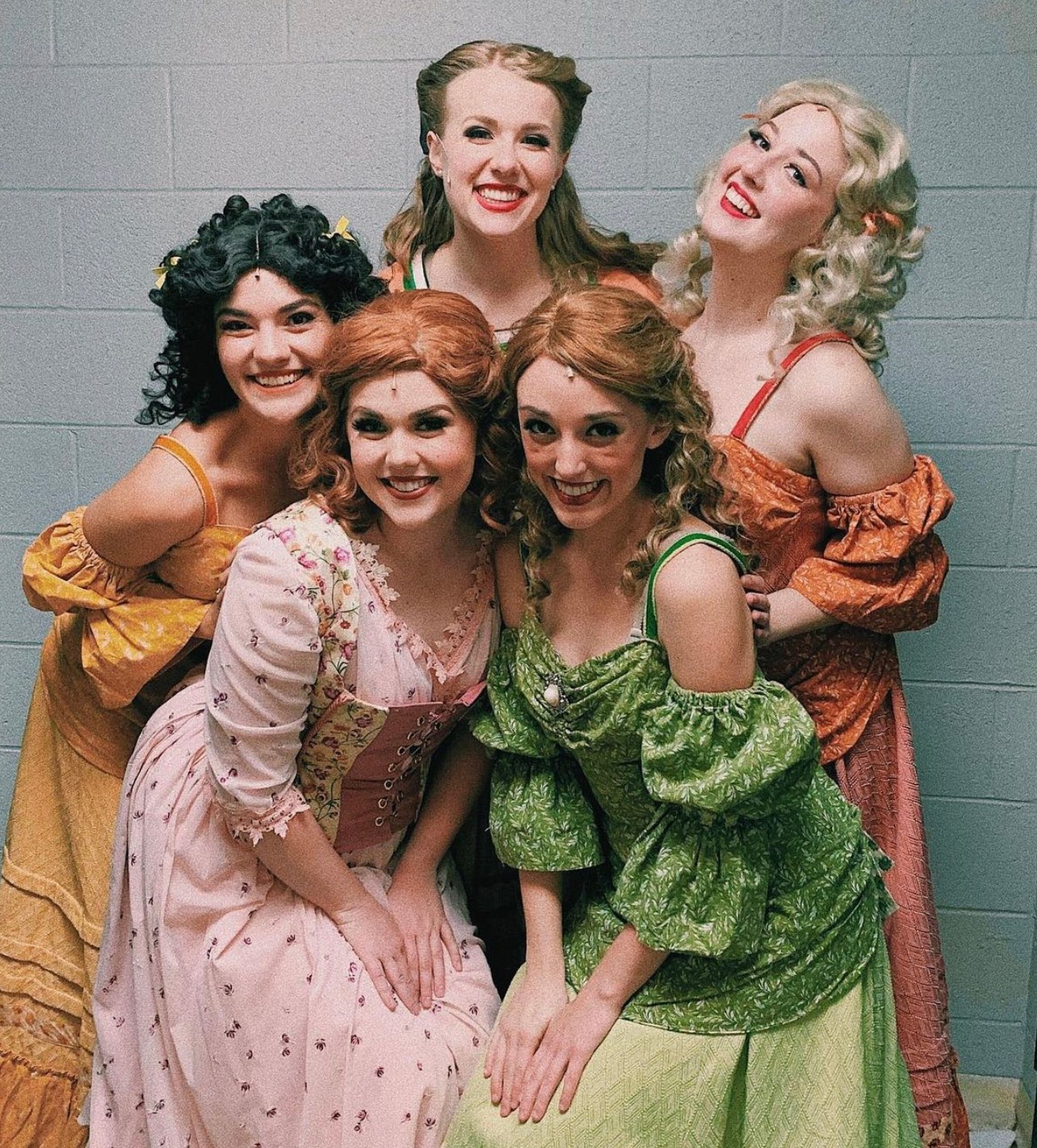 Five women in vintage dresses smiling, standing against a plain wall, with hairstyles and makeup reminiscent of the 1940s or 1950s.