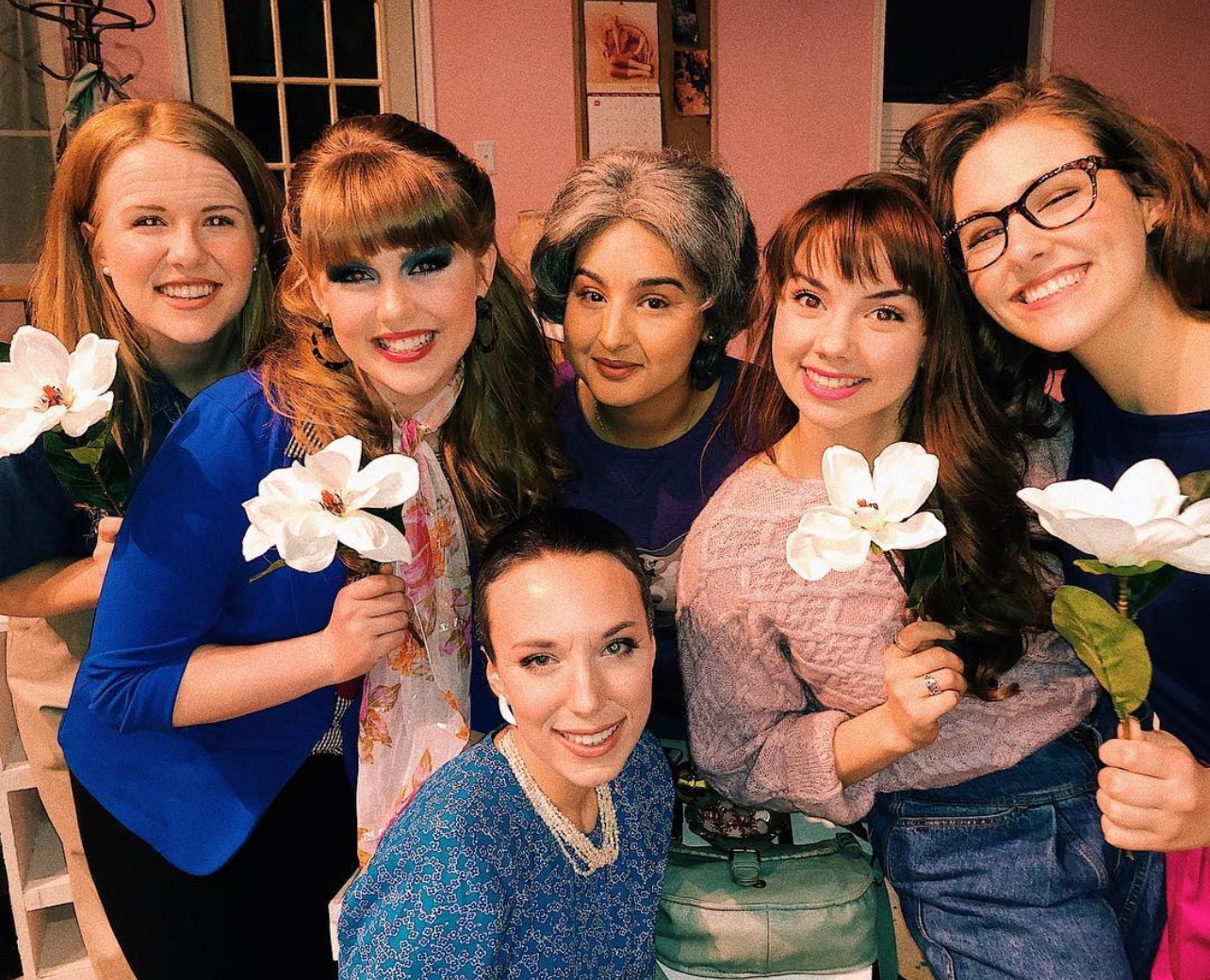 Group of five women smiling and holding white flowers indoors, with pink walls and framed pictures in the background.