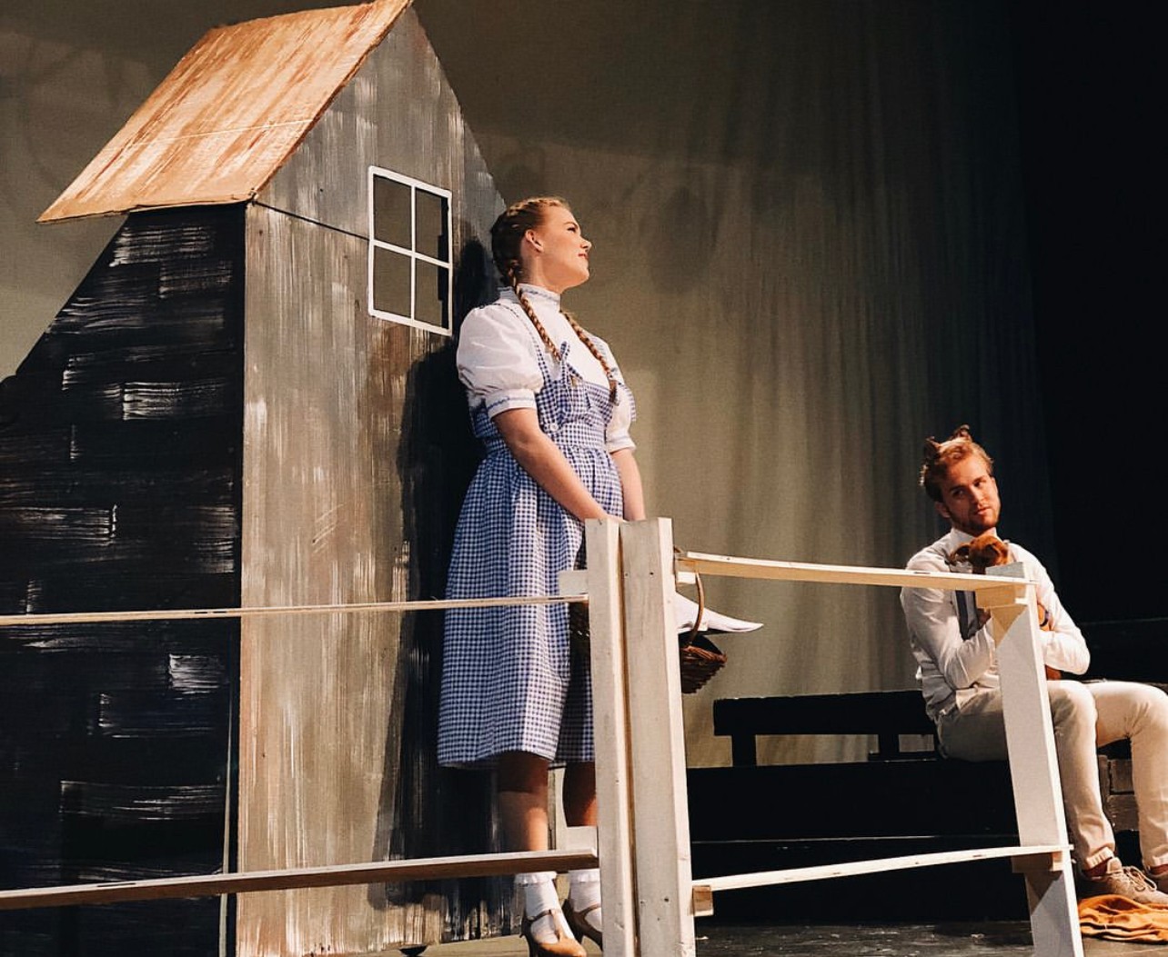 Two actors on stage, one standing and one sitting, with a small house prop in the background.