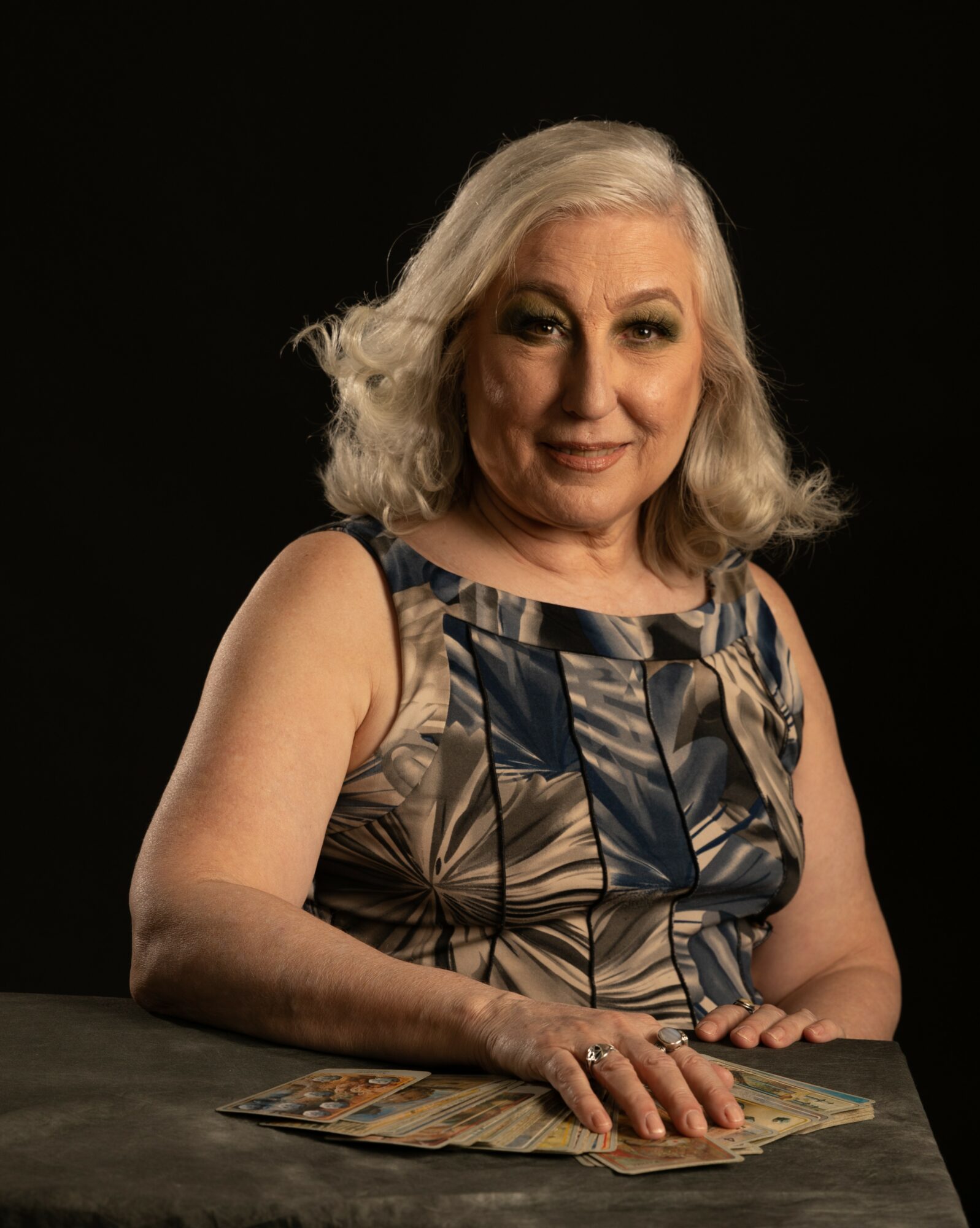 A woman with gray hair and a patterned sleeveless top sits at a table with a fan of paper in front of her, against a black background.