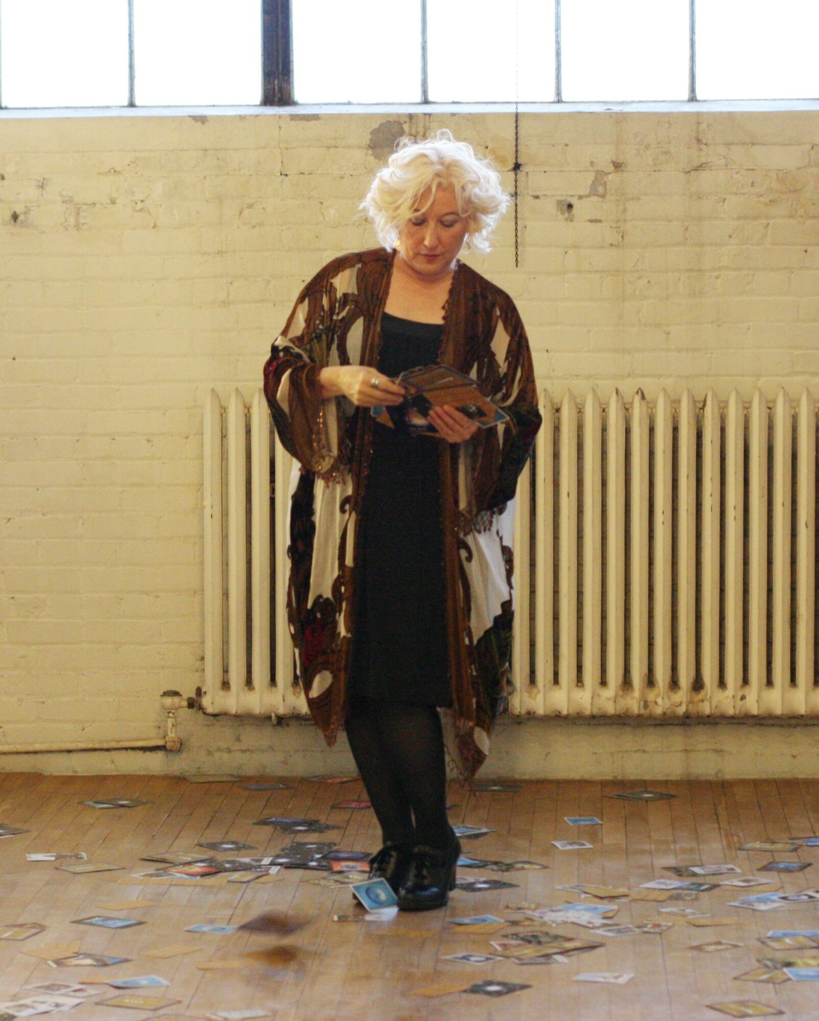 Older woman with curly white hair reads a magazine in a room with a brick wall and radiator, scattered papers on floor.