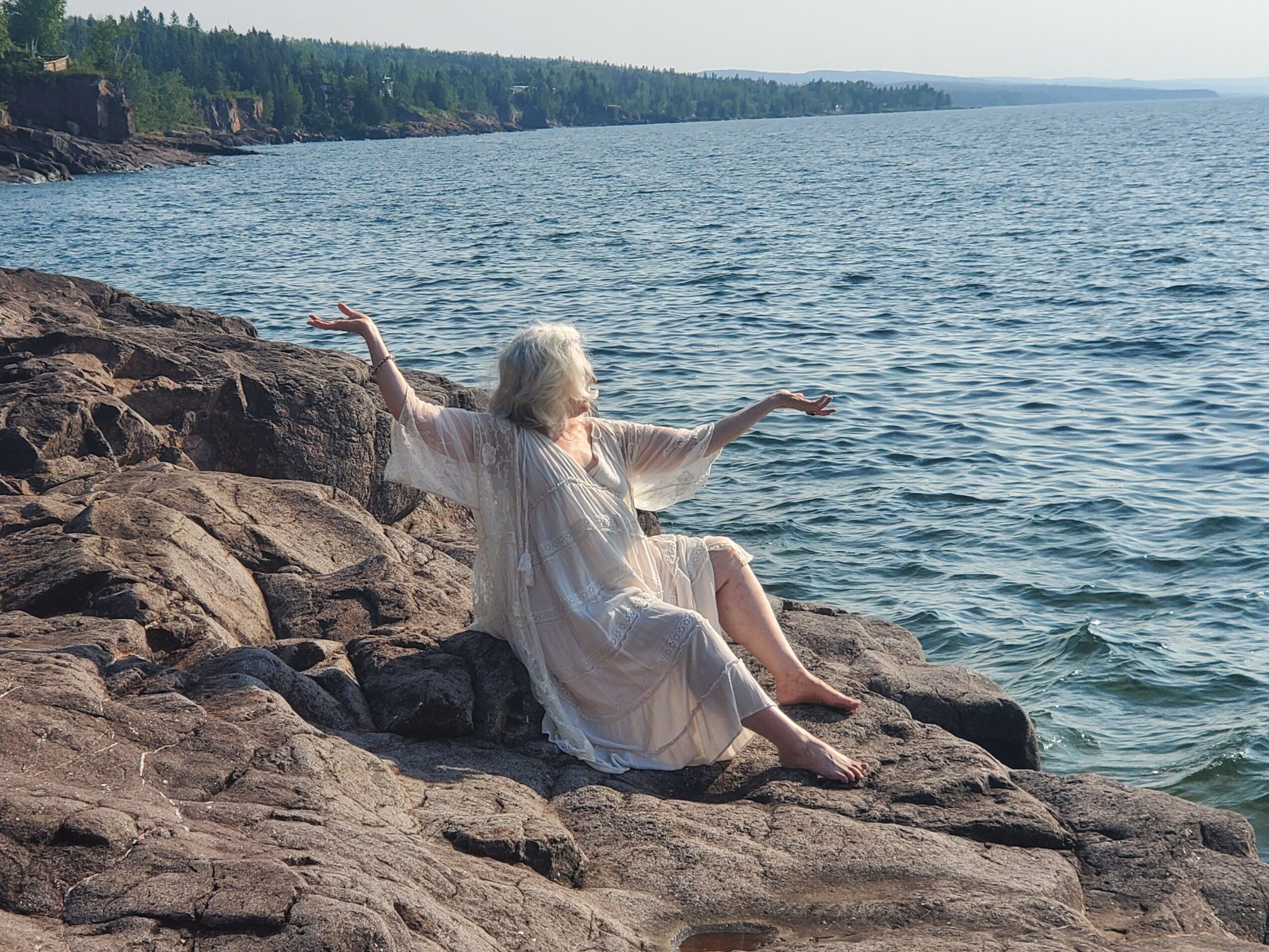 Woman in a white dress sitting on rocks by the water with arms outstretched, facing the sea, with a distant shoreline.