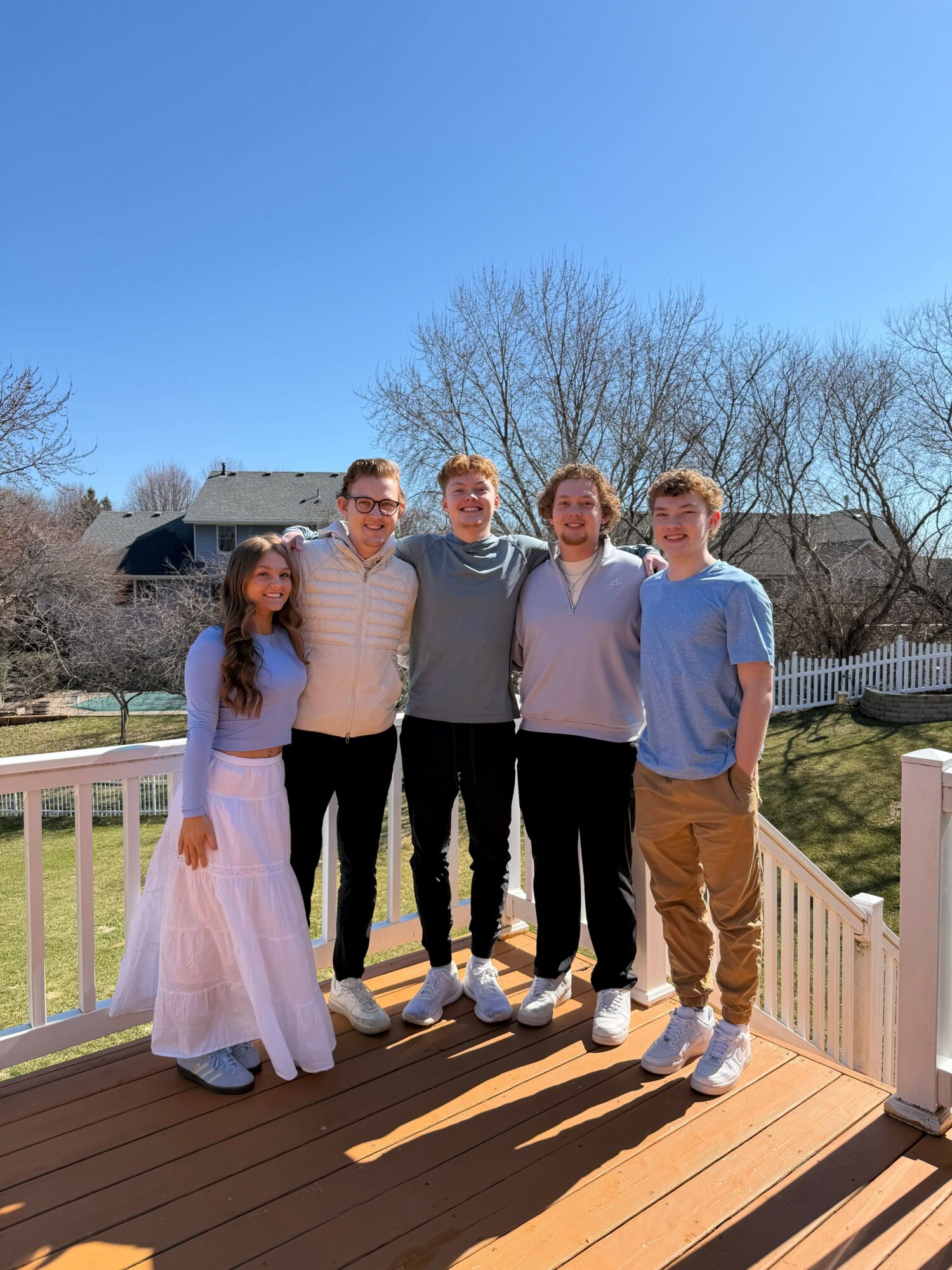 Four people standing on a wooden deck outdoors, with trees and a house in the background, under a clear blue sky.