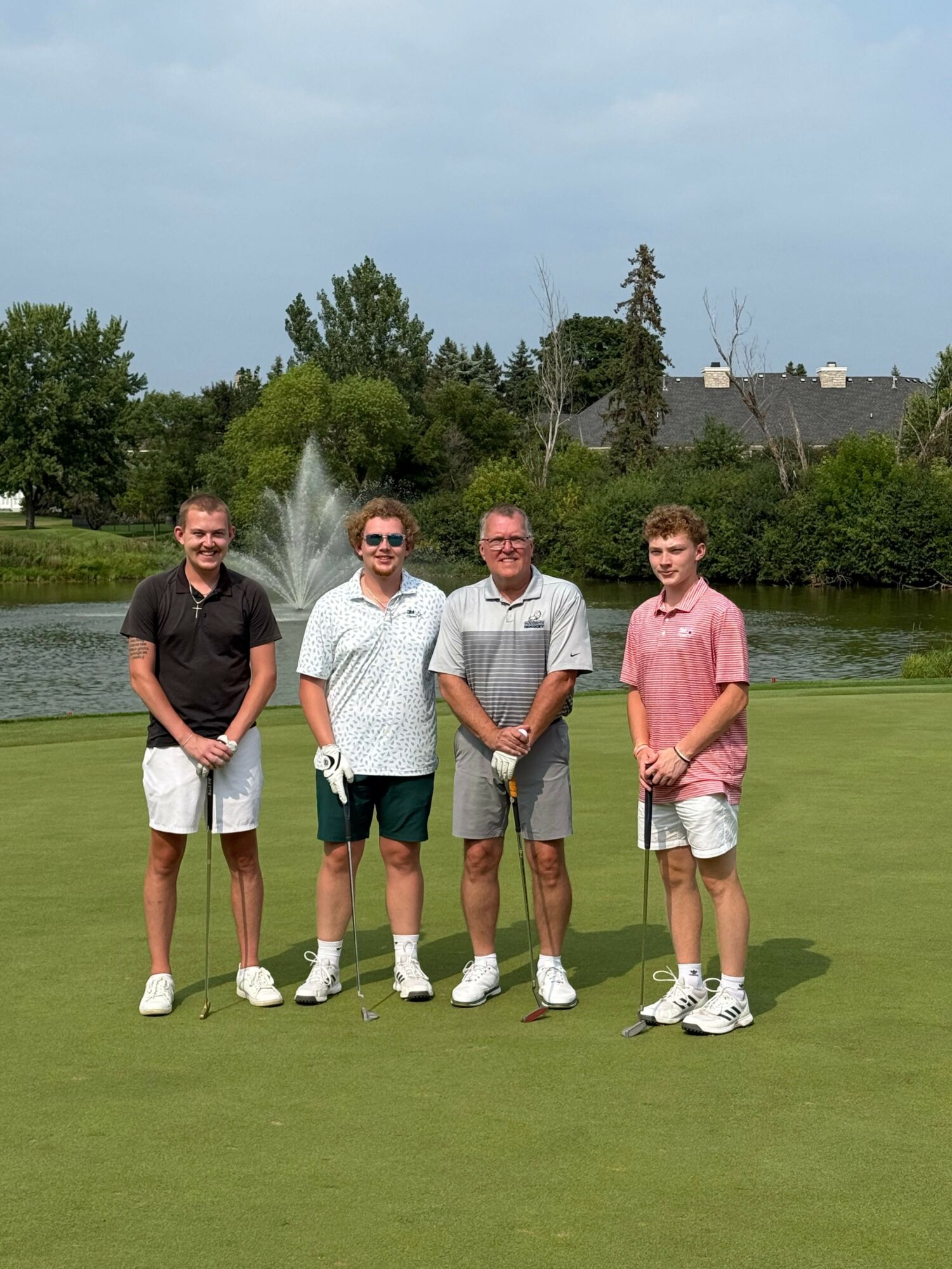Three people standing on a golf course with trees and buildings in the background, smiling at the camera.