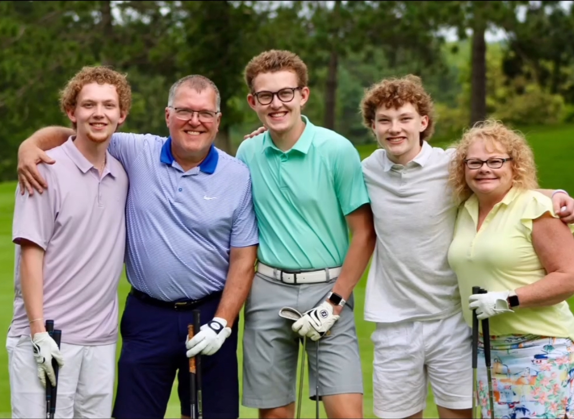 Group of five people smiling outdoors, holding golf clubs, with trees in the background.