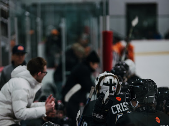People sitting and standing near a glass barrier at an indoor ice hockey rink, some wearing helmets and jackets.