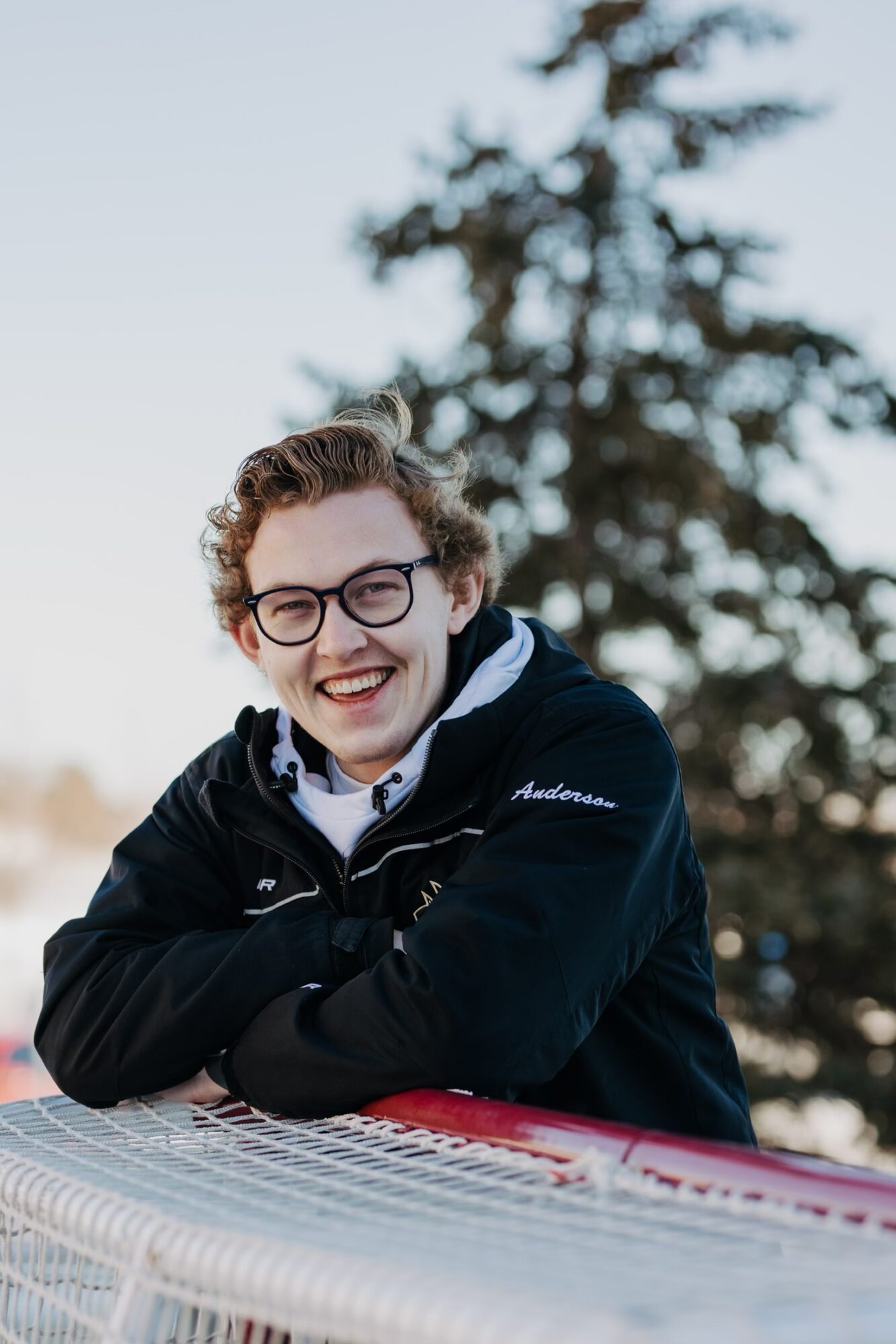 Person with glasses smiling outdoors, leaning on a surface, with a tree in the background.