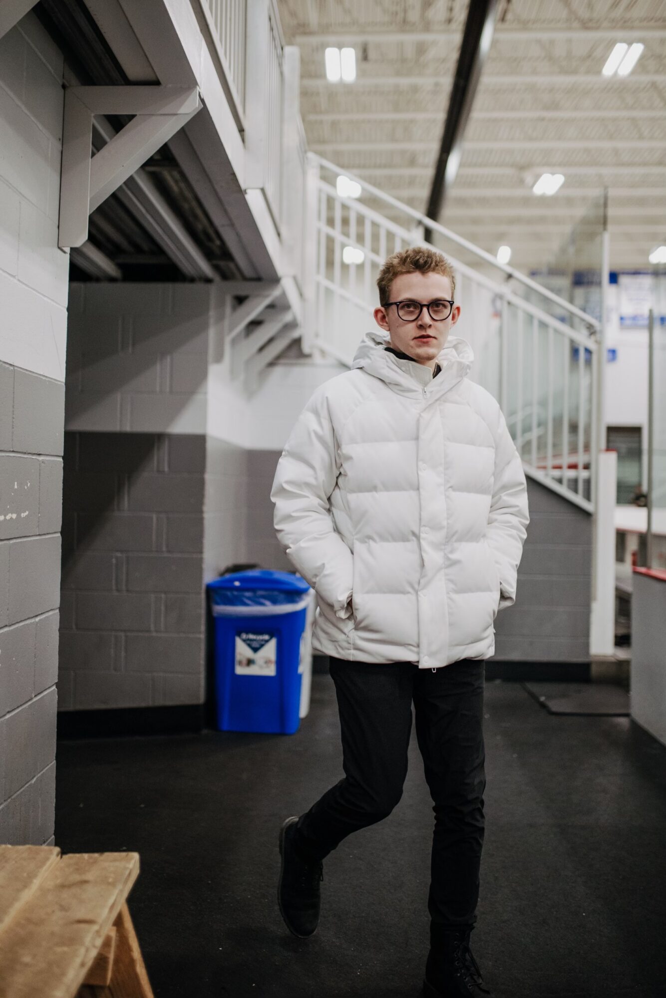 Young person in white puffer jacket and glasses walking indoors, with blue trash bin and staircase in background.