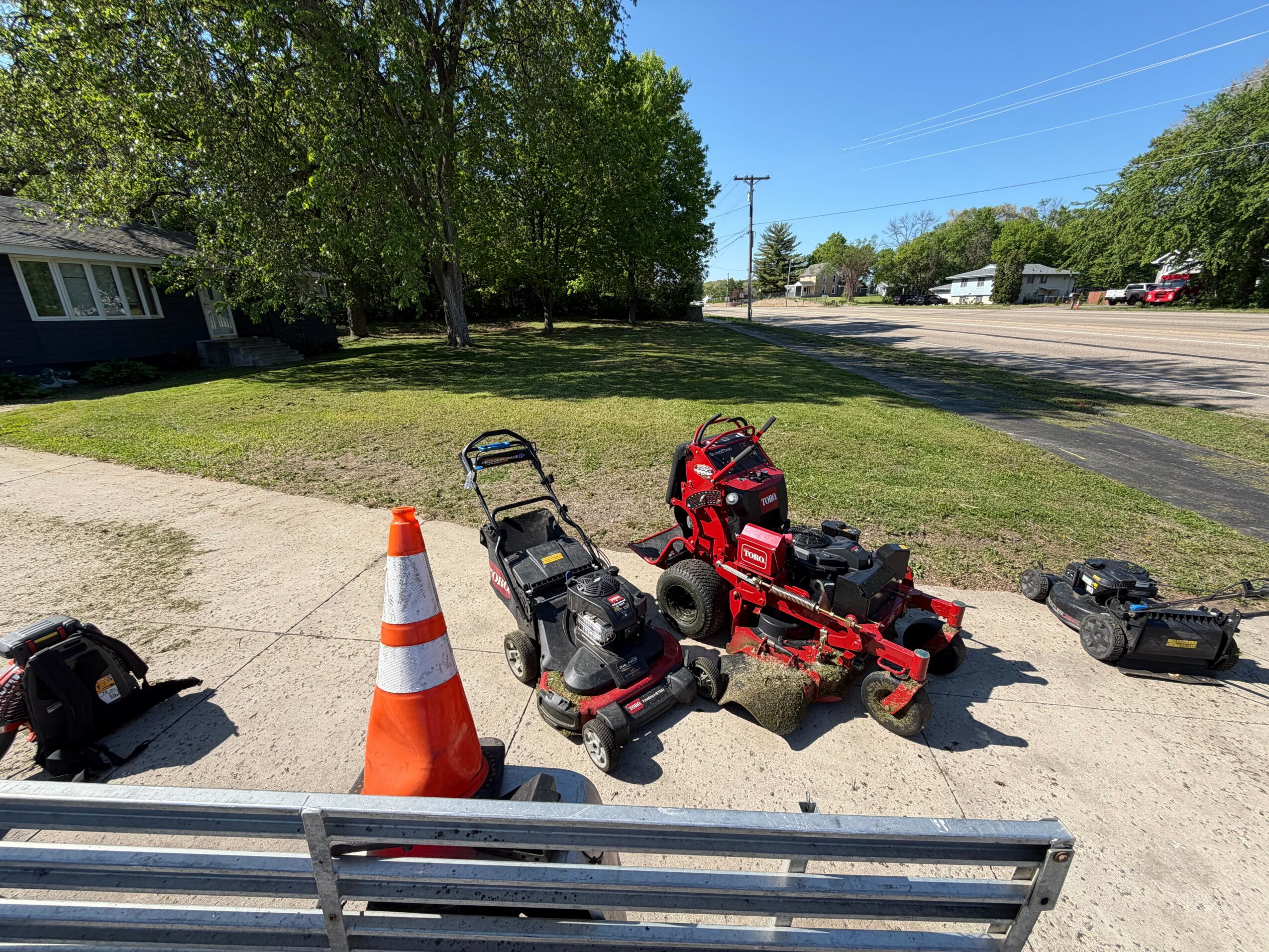 Lawnmowers and a traffic cone on a sidewalk with grass, trees, and a street in the background.