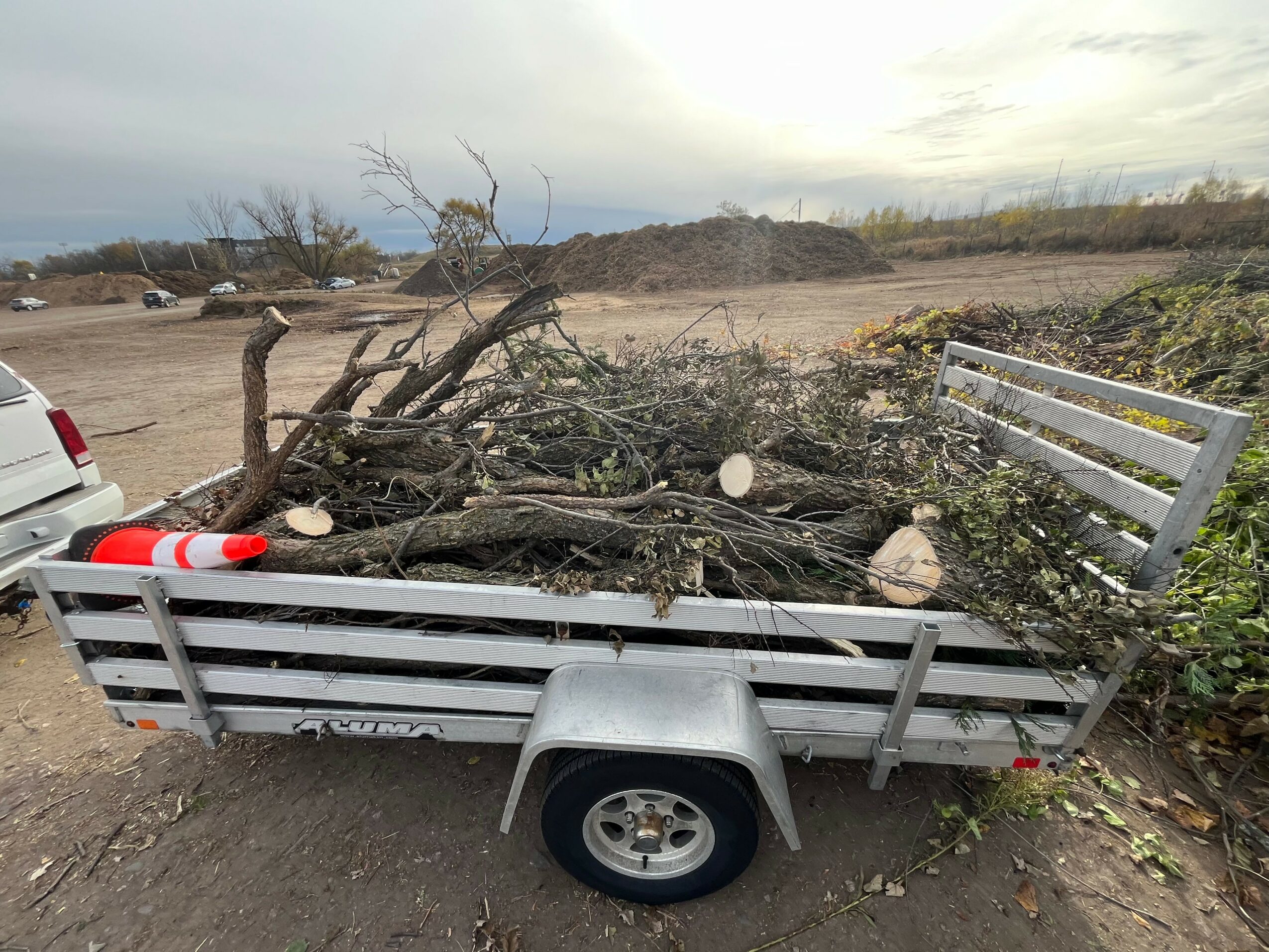 Trailer filled with fallen branches and debris, parked outdoors with a cloudy sky in the background.