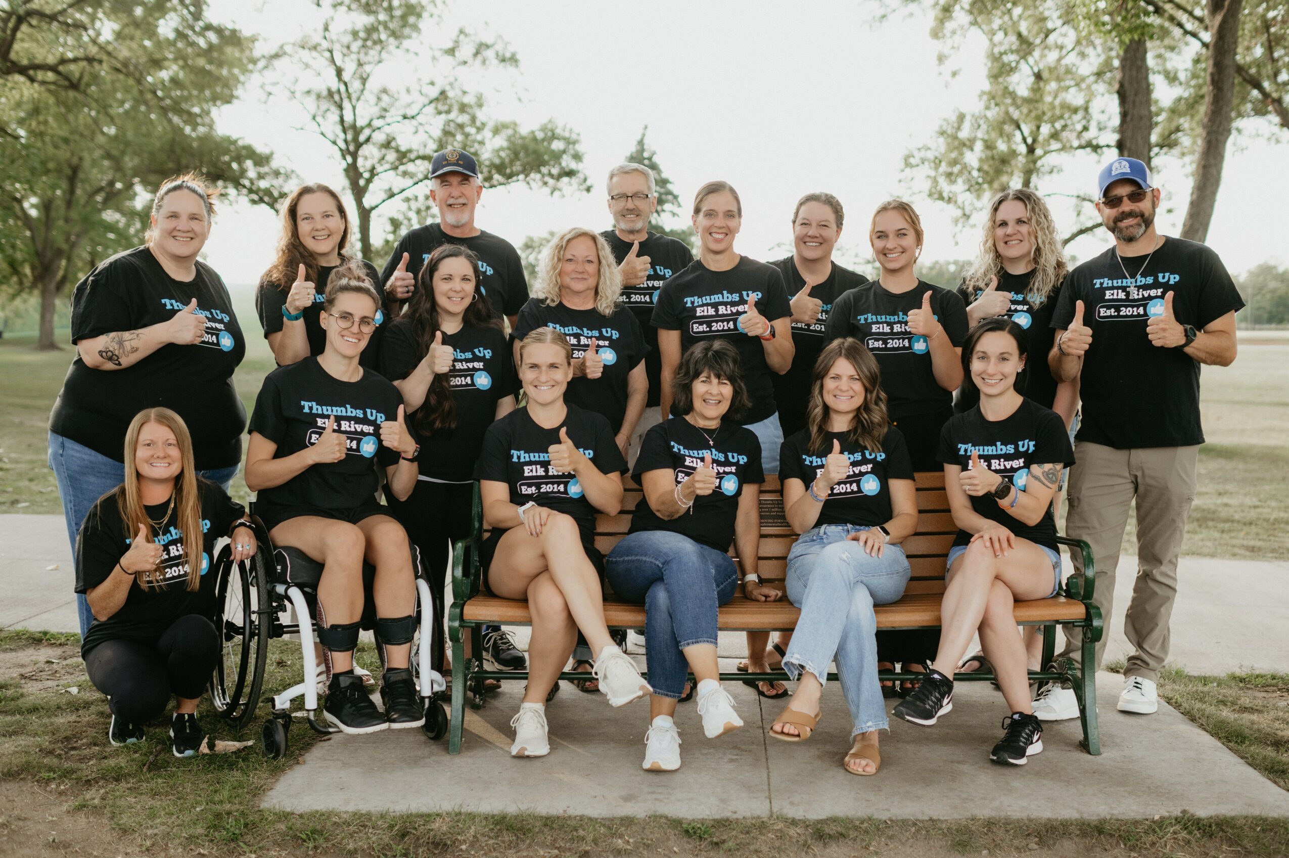 Group of 16 people outdoors, some in wheelchairs, wearing matching black T-shirts, smiling, with trees and water in background.