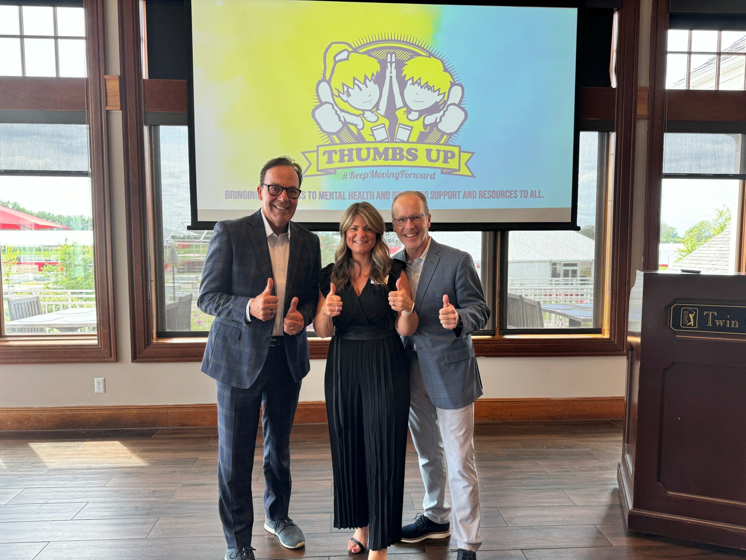 Three people standing indoors in front of a large screen, giving thumbs up, smiling, with windows behind them.
