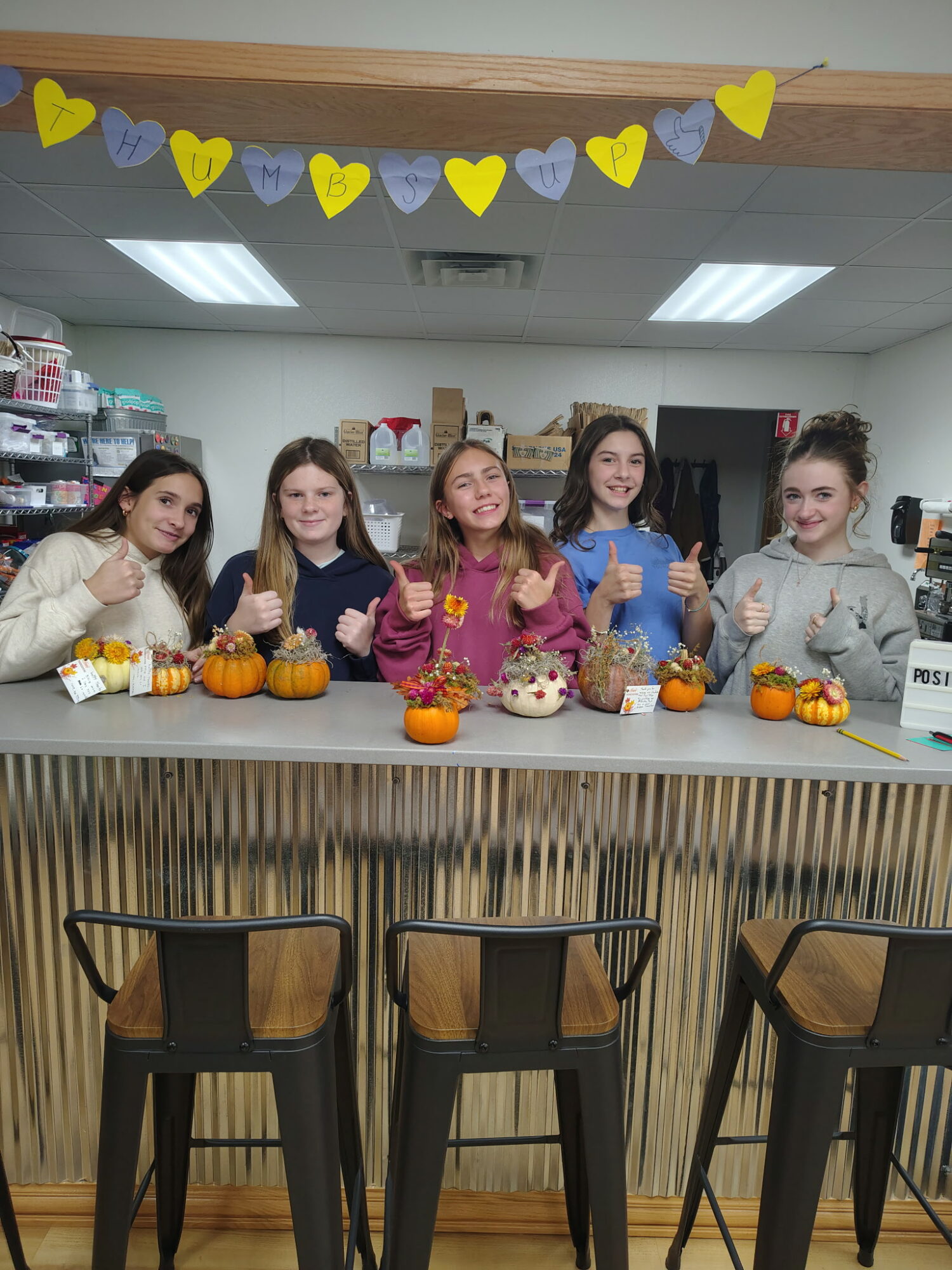 Five young girls stand behind a counter with small pumpkins and baked goods, smiling and giving thumbs up, in a decorated room.