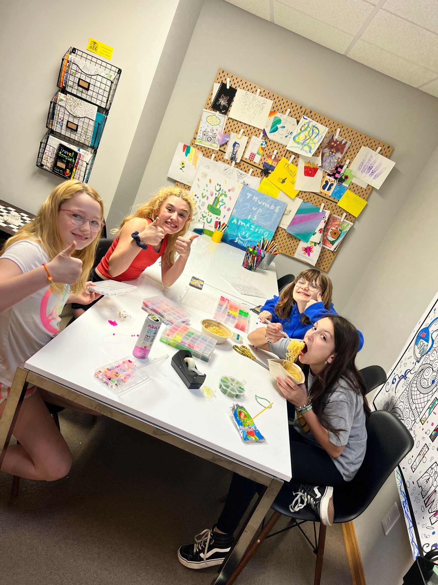Four children sitting around a table with craft supplies, smiling and looking at the camera, in a classroom setting.
