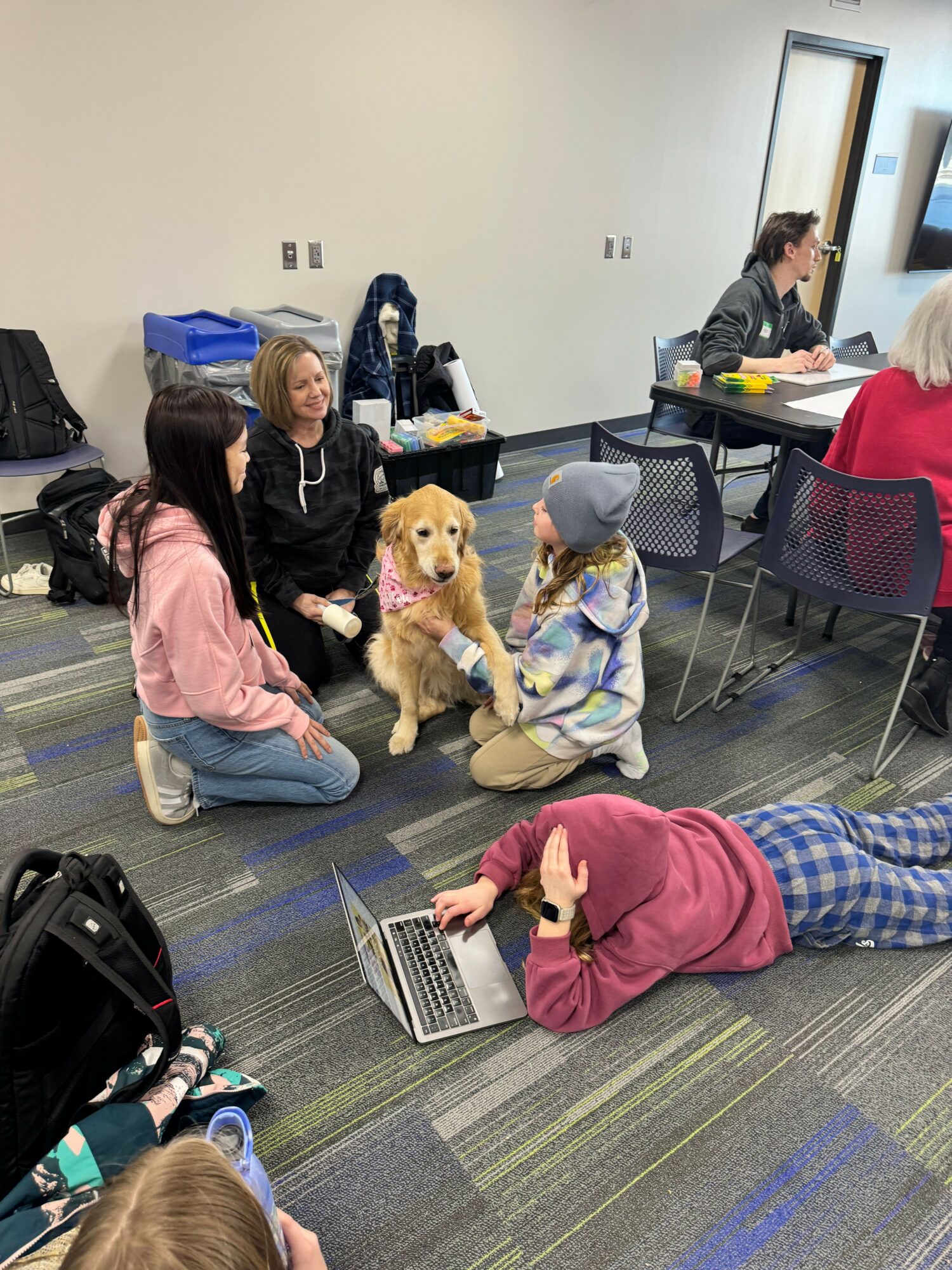 People sitting on chairs and floor in a room, some using laptops, with a dog among them, and a person lying on the floor.