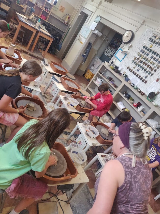Children and an adult working on pottery projects in a classroom with shelves and tools, some children wearing aprons.