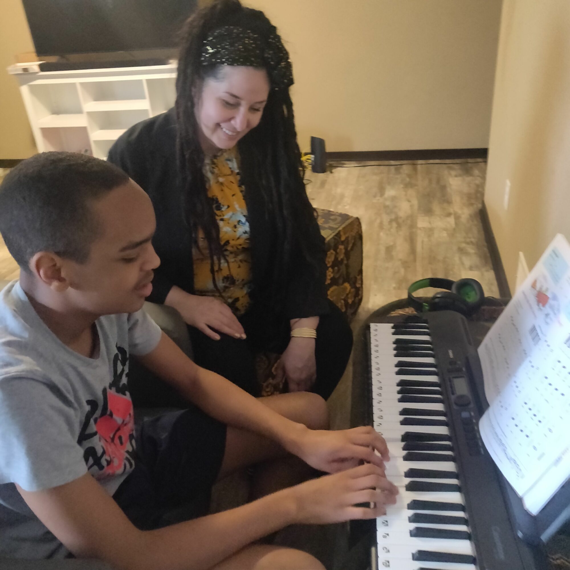 Two children sit at a piano, one playing and the other watching, in a room with a beige wall and wooden floor.