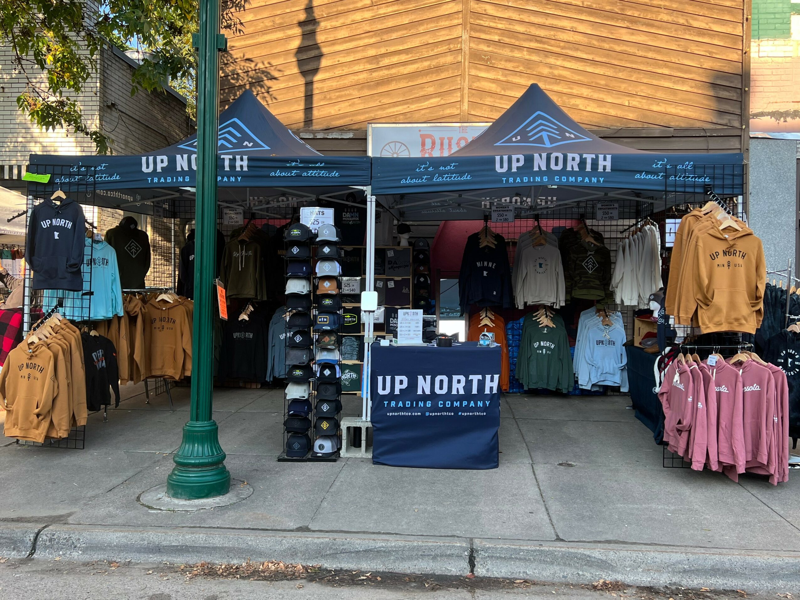 Outdoor clothing stall with hats, jackets, and shirts displayed under tents on a sidewalk.