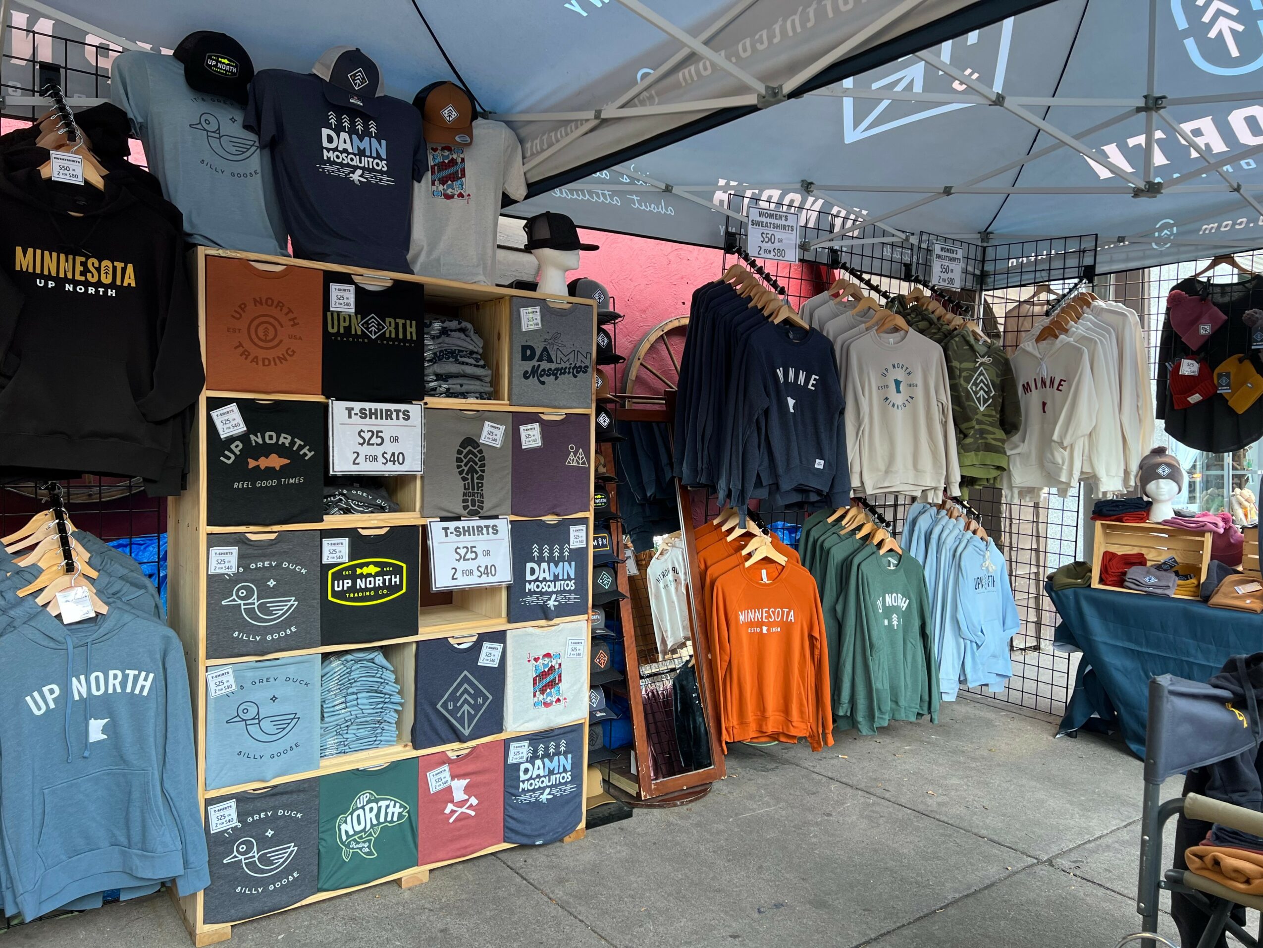 Clothing display at an outdoor market stall with shirts, hoodies, and boxes of merchandise under a tent.