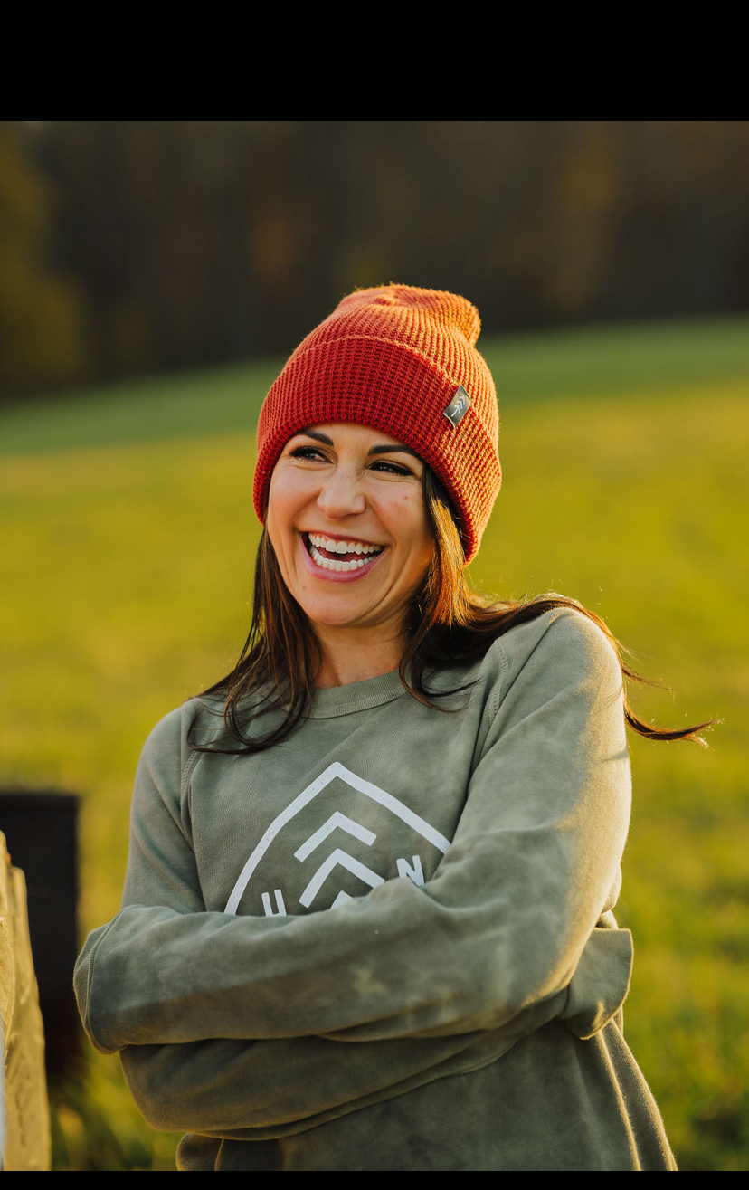 Woman smiling outdoors wearing a red beanie and green sweatshirt, with a grassy field in the background.