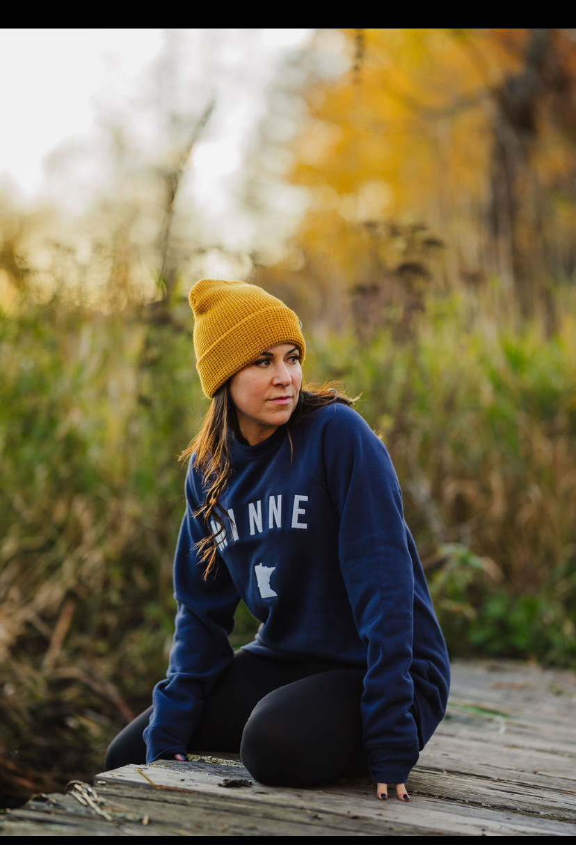 Young woman sitting outdoors on a wooden path, wearing a yellow beanie and navy sweater, with autumn trees in background.