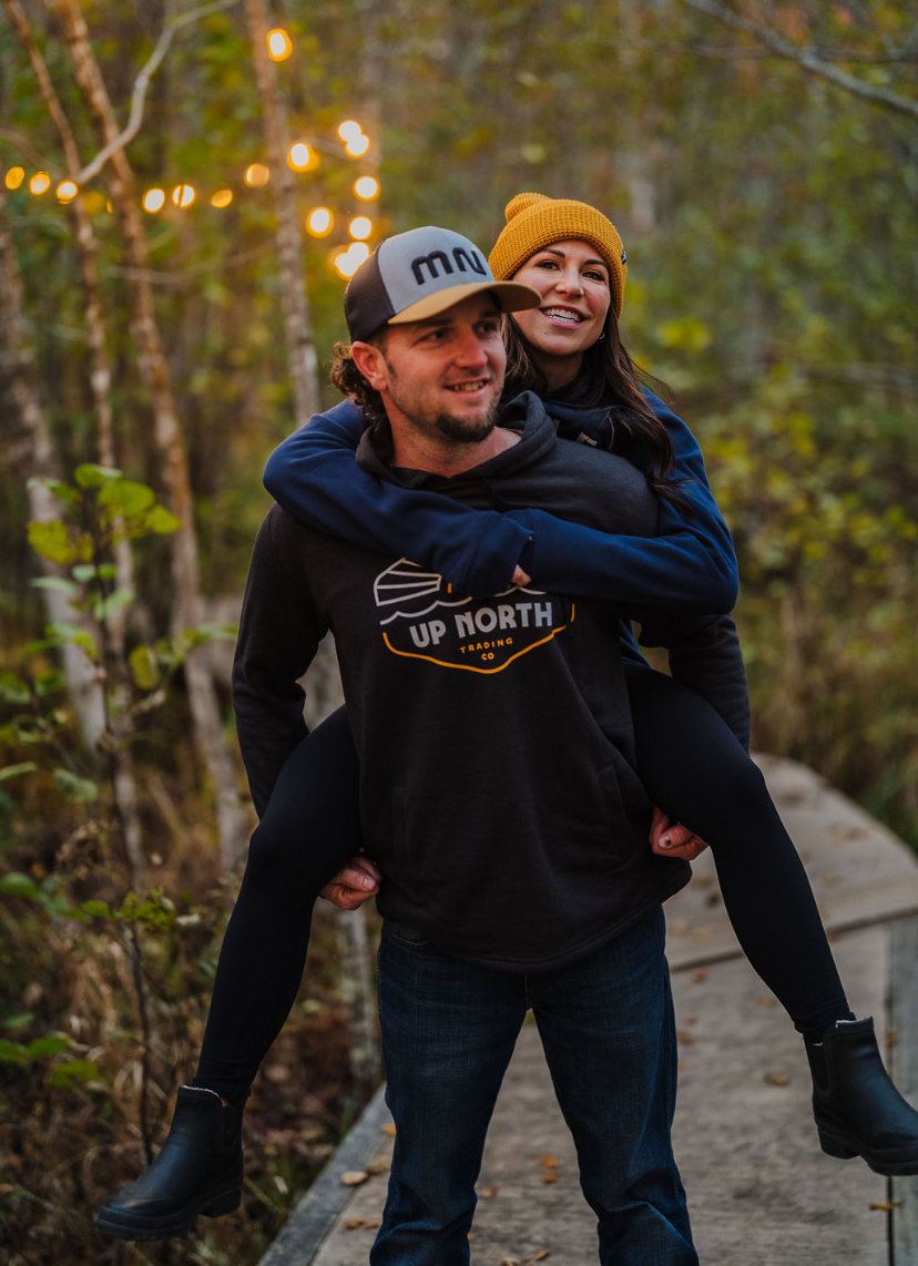 A man carrying a woman on his back outdoors in a wooded area with string lights, both smiling, wearing casual clothing.
