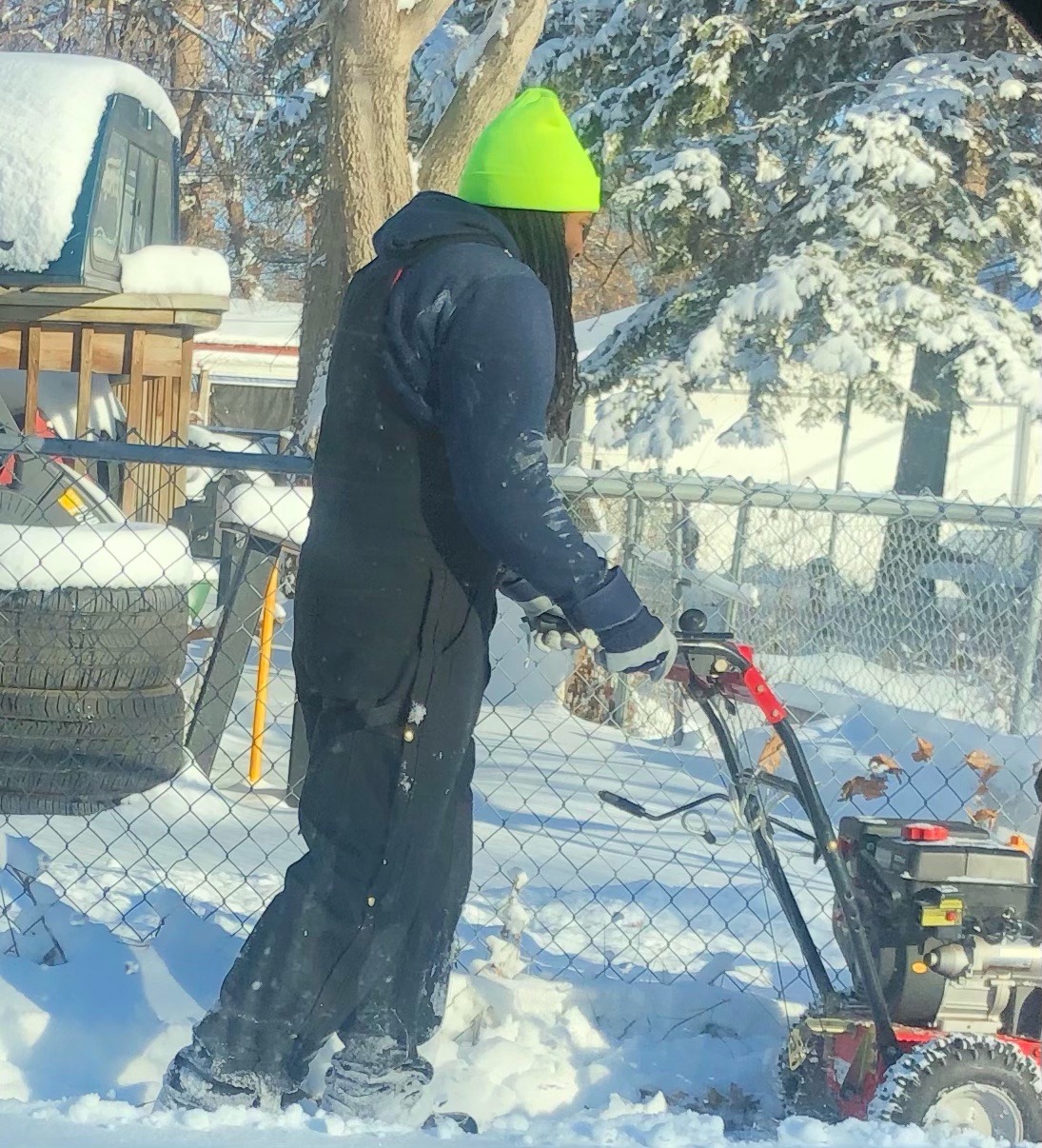 Person in winter clothing using a snow blower outdoors with snow-covered trees and fence in background.