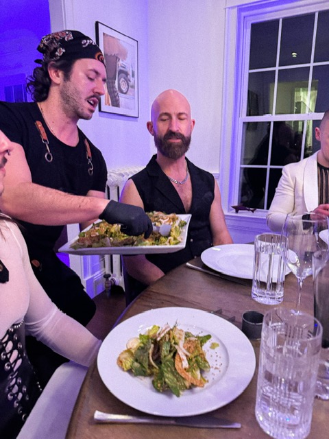 Person serving food to two seated men at a dinner table, with a plate of salad in foreground, window in background.
