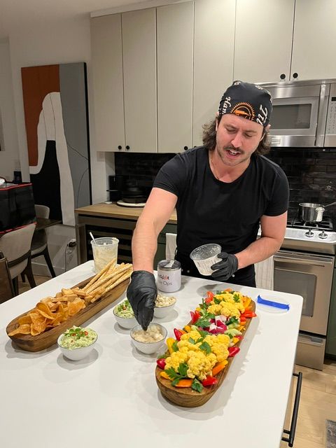 Person wearing a black shirt and cap, smiling, preparing a colorful cake with yellow, red, and green decorations on a white table.