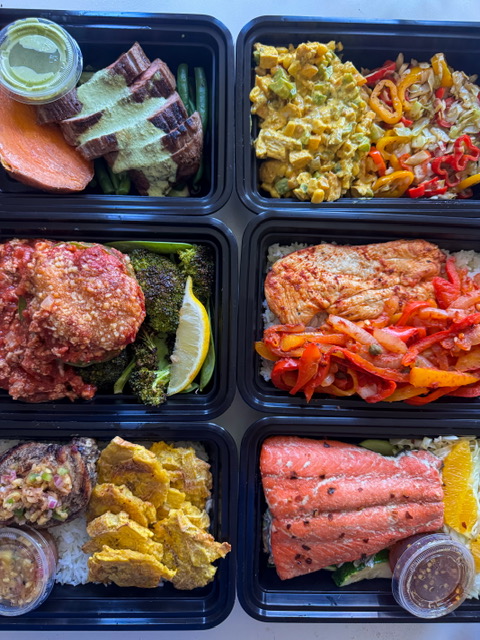 Six black meal containers with various foods including salmon, chicken, vegetables, and sides, arranged in two rows of three.