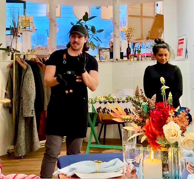 Two people standing inside a store with clothing and flowers, one wearing a black apron and cap, the other in black clothing.